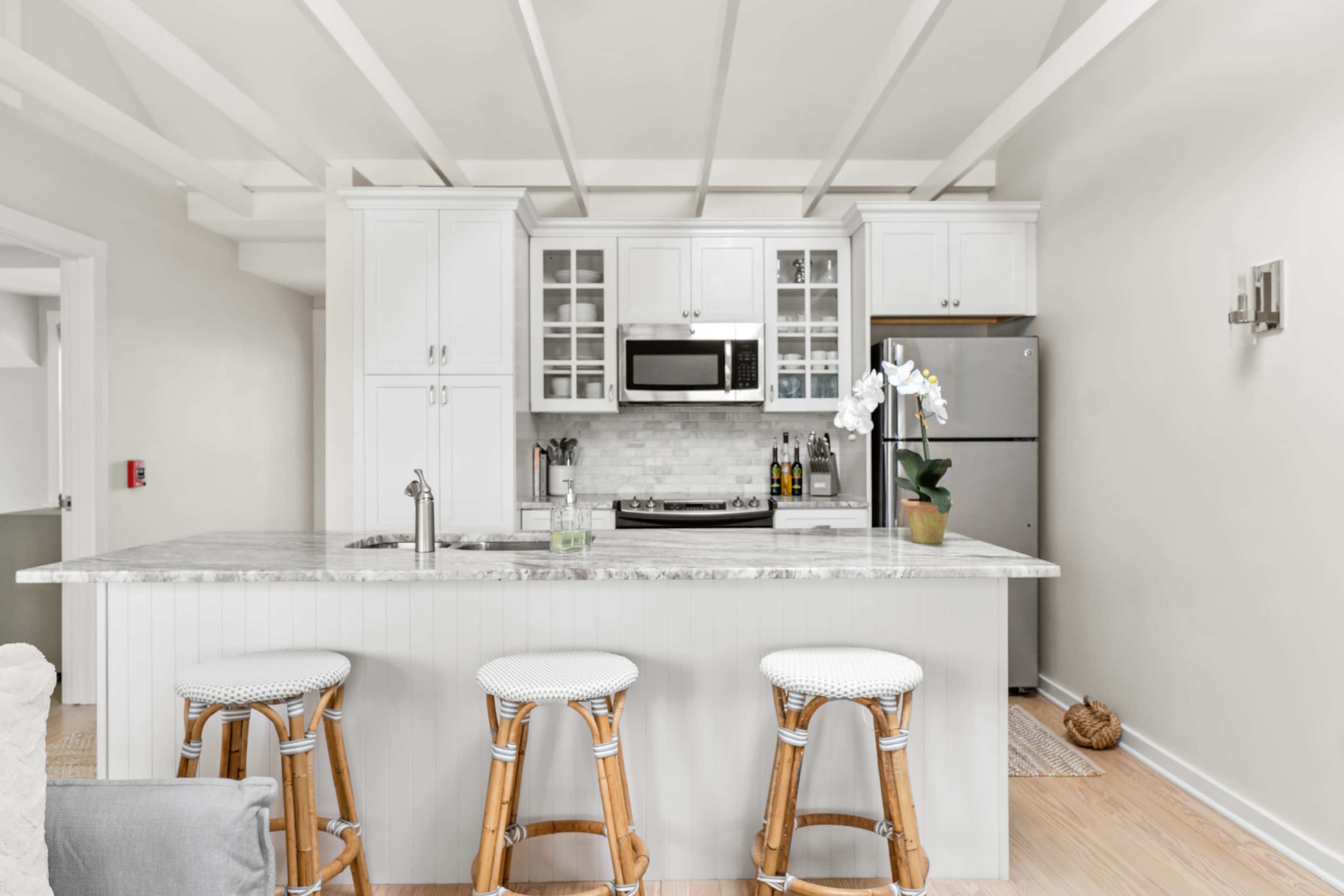 A modern kitchen features a marble countertop with three woven stools, white cabinetry, and stainless steel appliances.