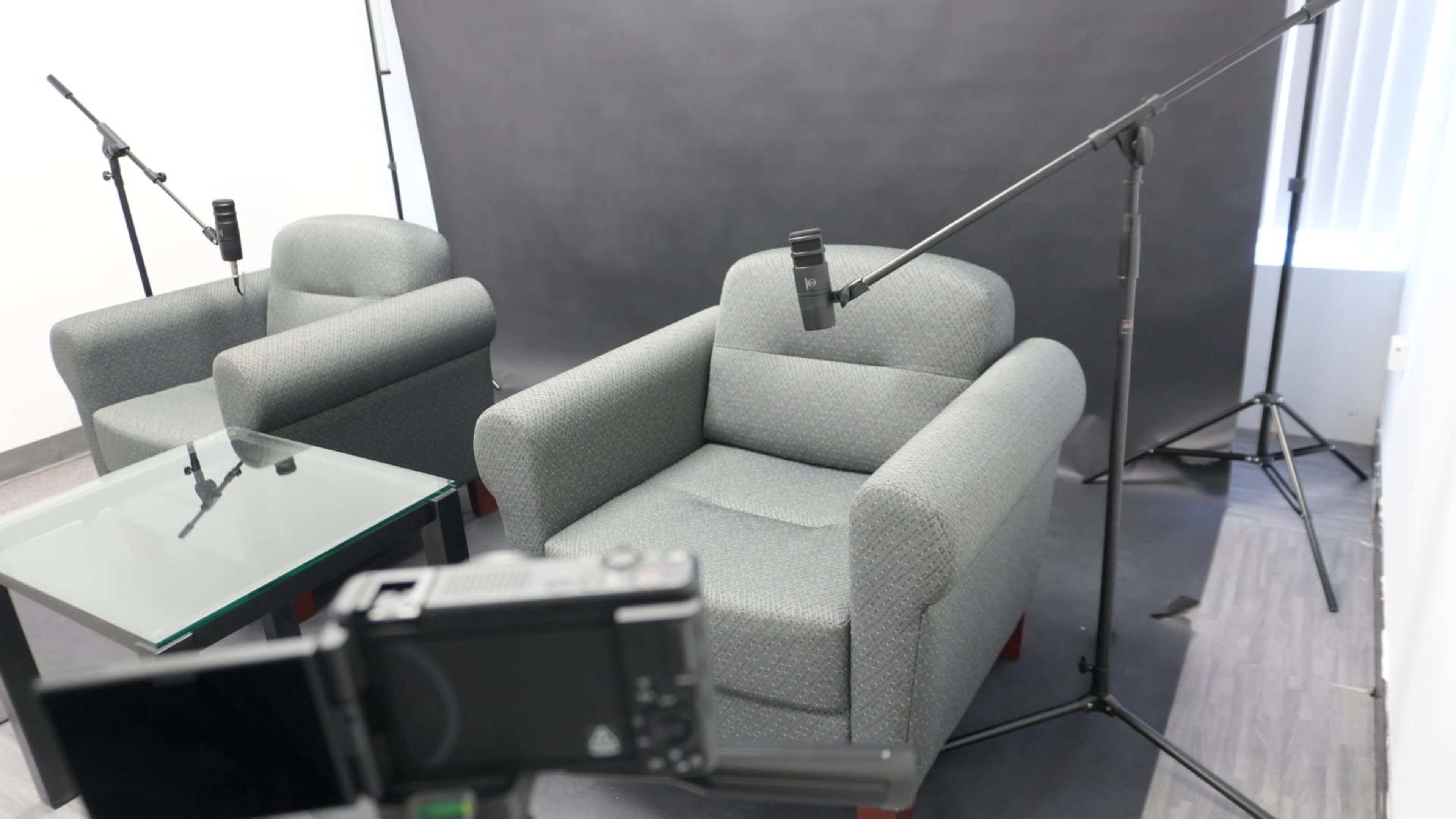 The image shows two gray armchairs facing each other with a glass table in between, and microphones mounted on stands in a room with a dark backdrop.
