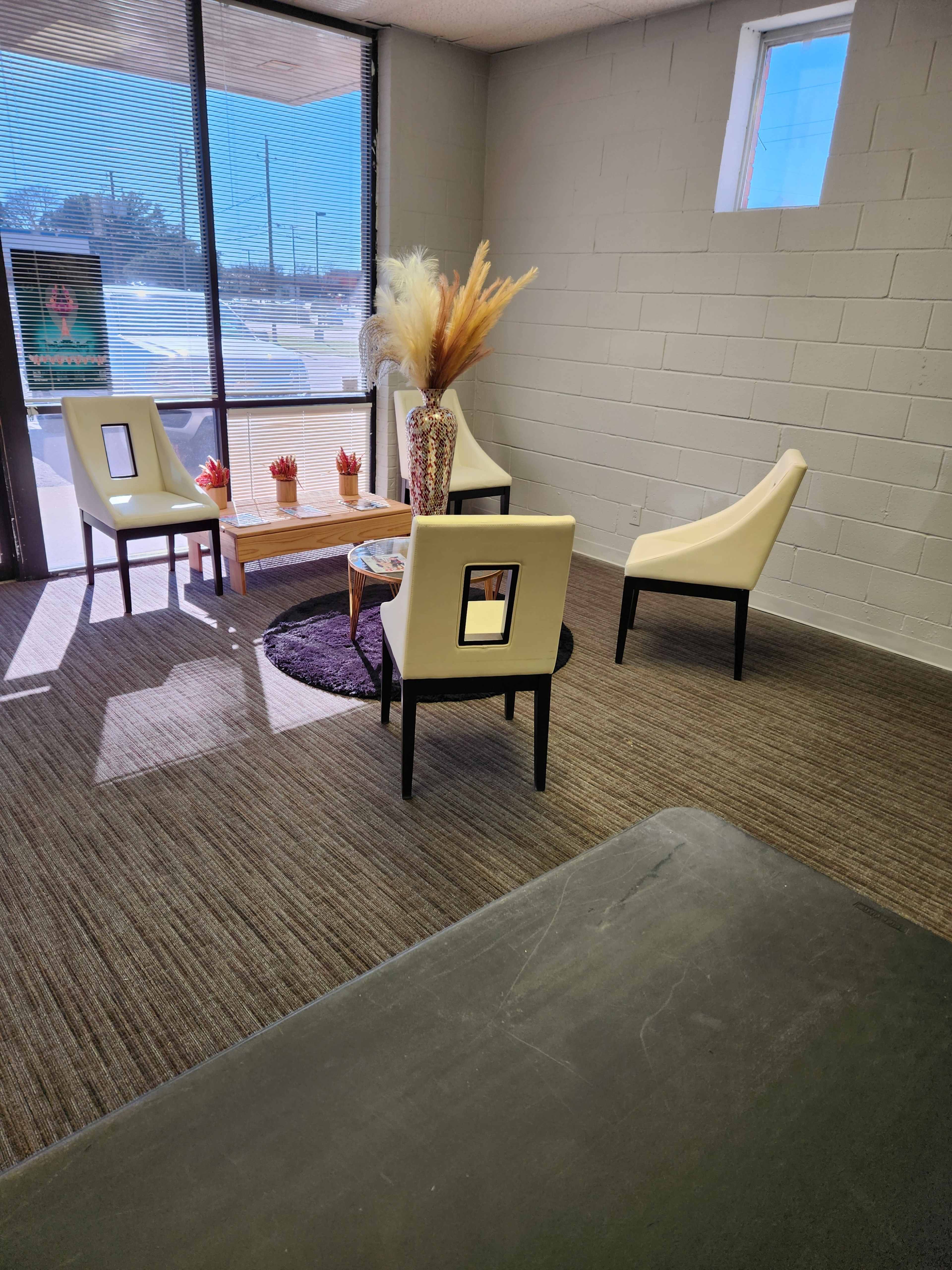 A small waiting area with two white chairs, a wooden coffee table, and a decorative vase with dried flowers, illuminated by natural light from a large window.