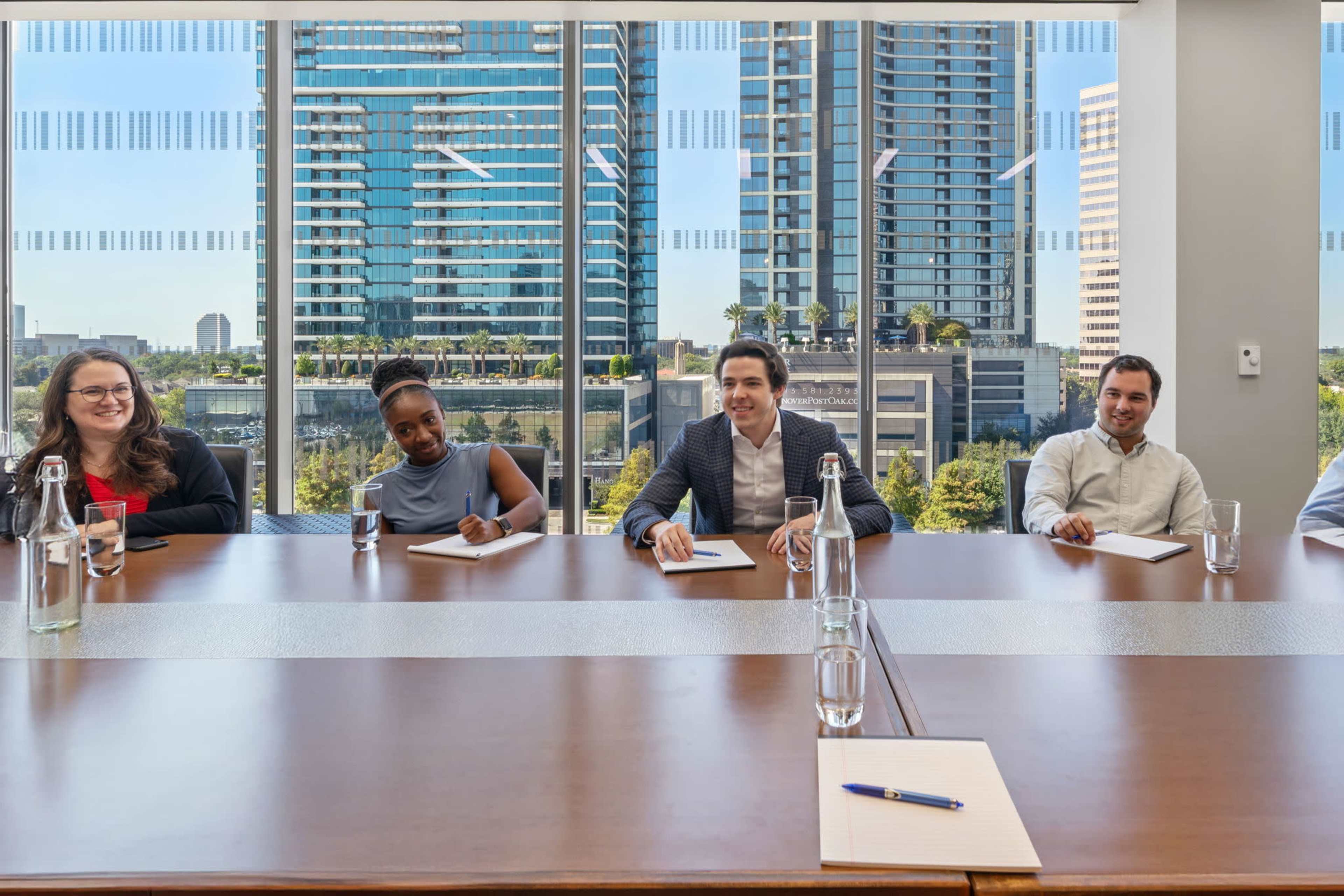 A group of five professionals sit at a conference table, engaged in a meeting with a view of tall buildings through large windows.