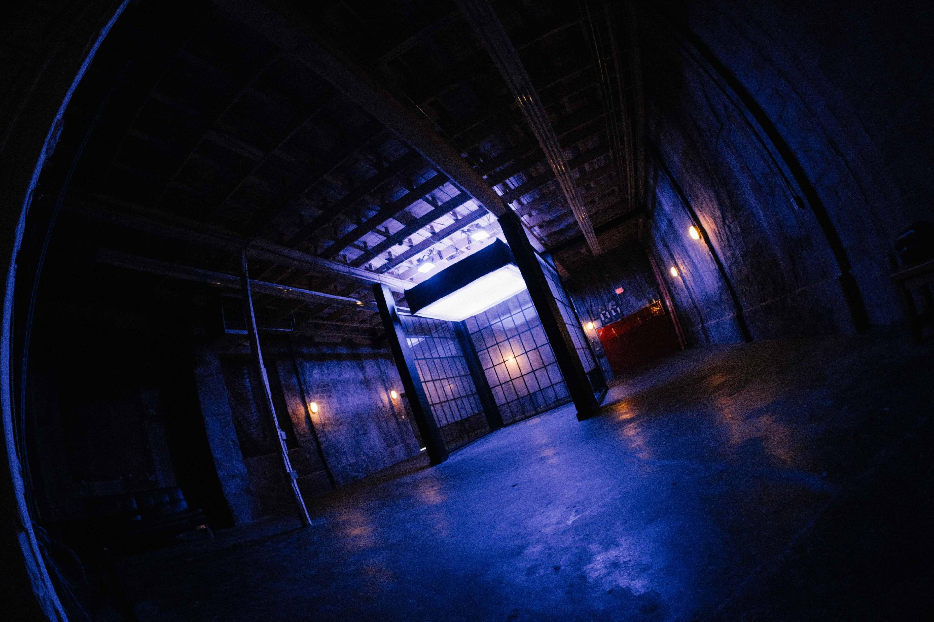 The image shows a large, dimly lit industrial space featuring a grid of beams overhead and a set of glass doors illuminated by a soft light.