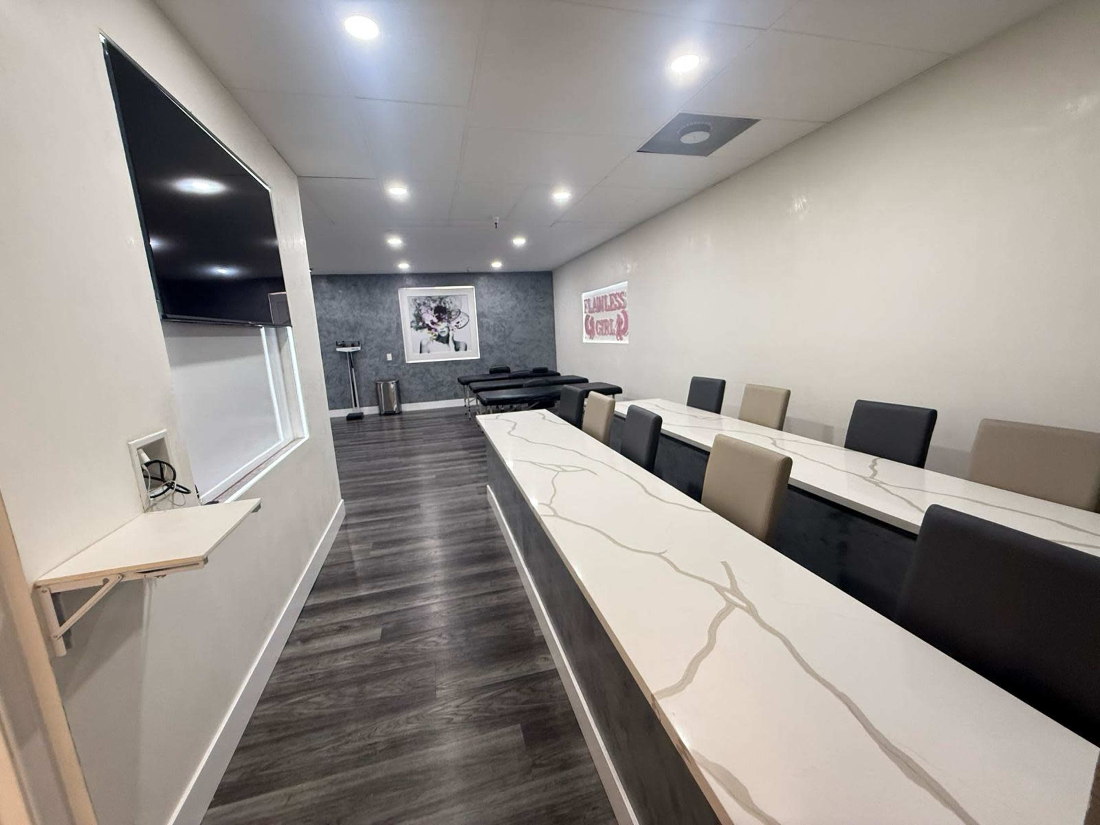 The image shows a modern meeting room with a long white table, chairs arranged on both sides, a wall-mounted screen, and simple decor.