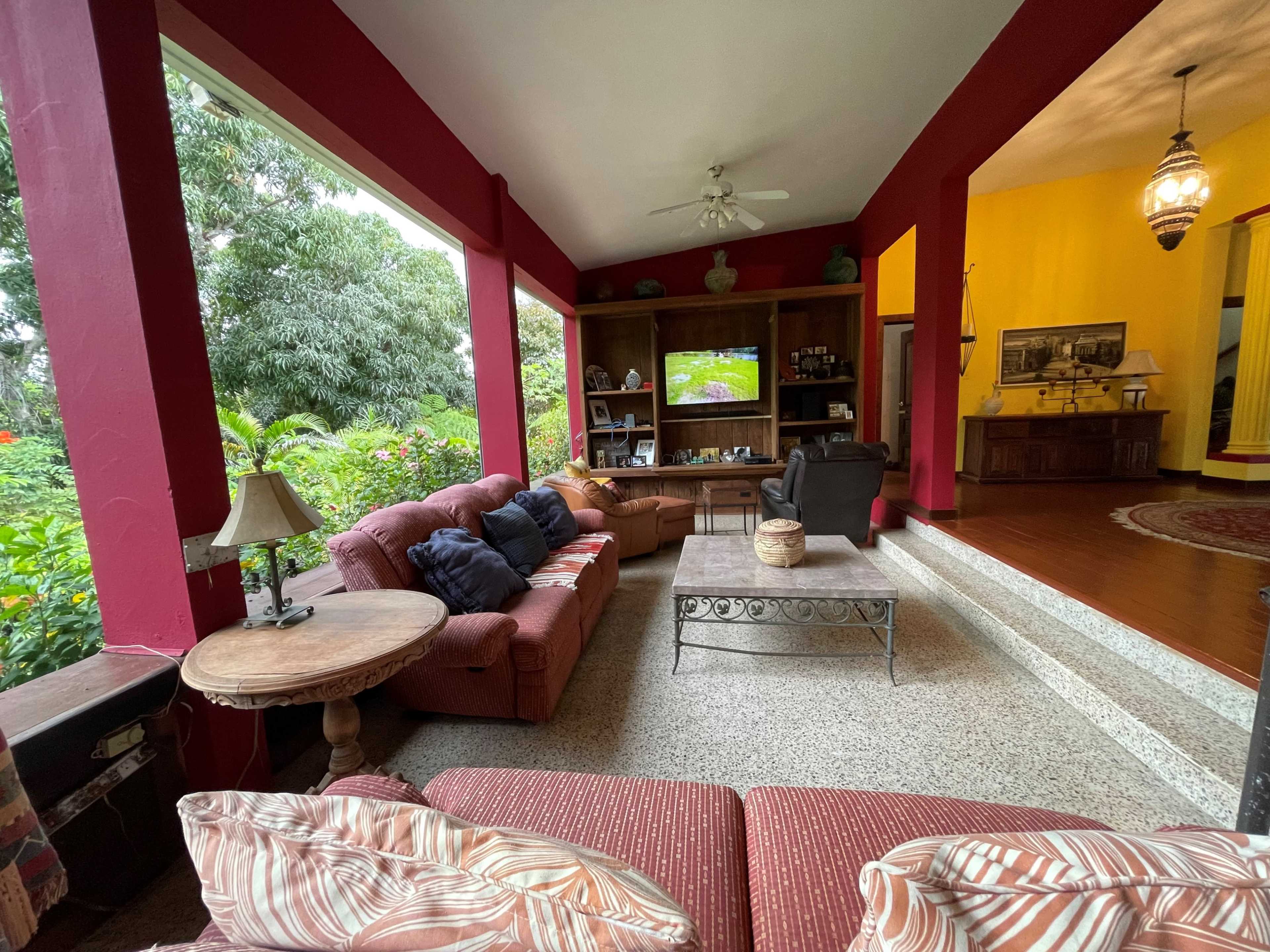 A living room with red walls, a seating area featuring a sofa and armchair, and a large window overlooking a lush garden.