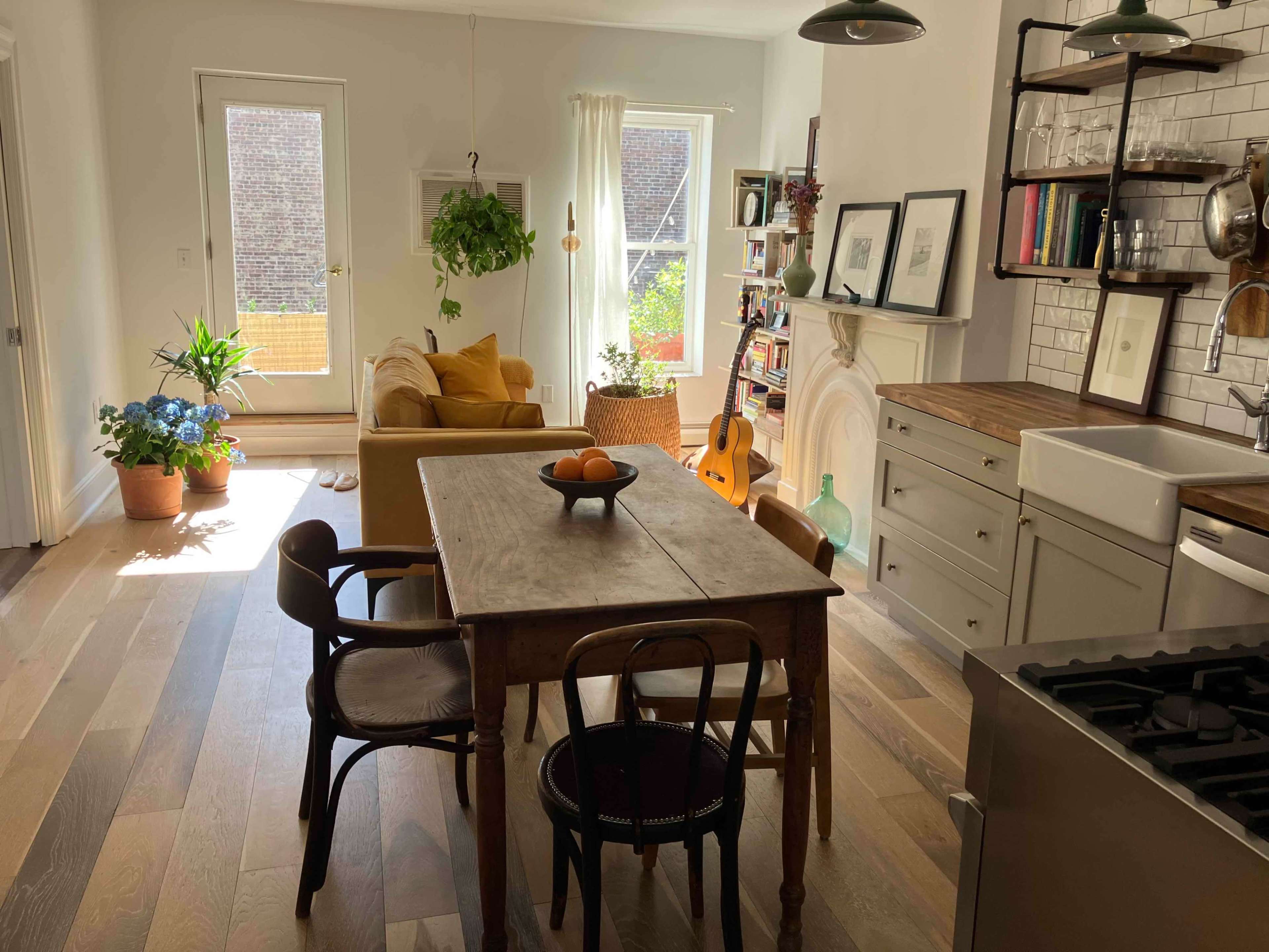 A bright kitchen and dining area features a wooden table, chairs, a yellow sofa, a guitar, and plants, with large windows allowing natural light to enter.