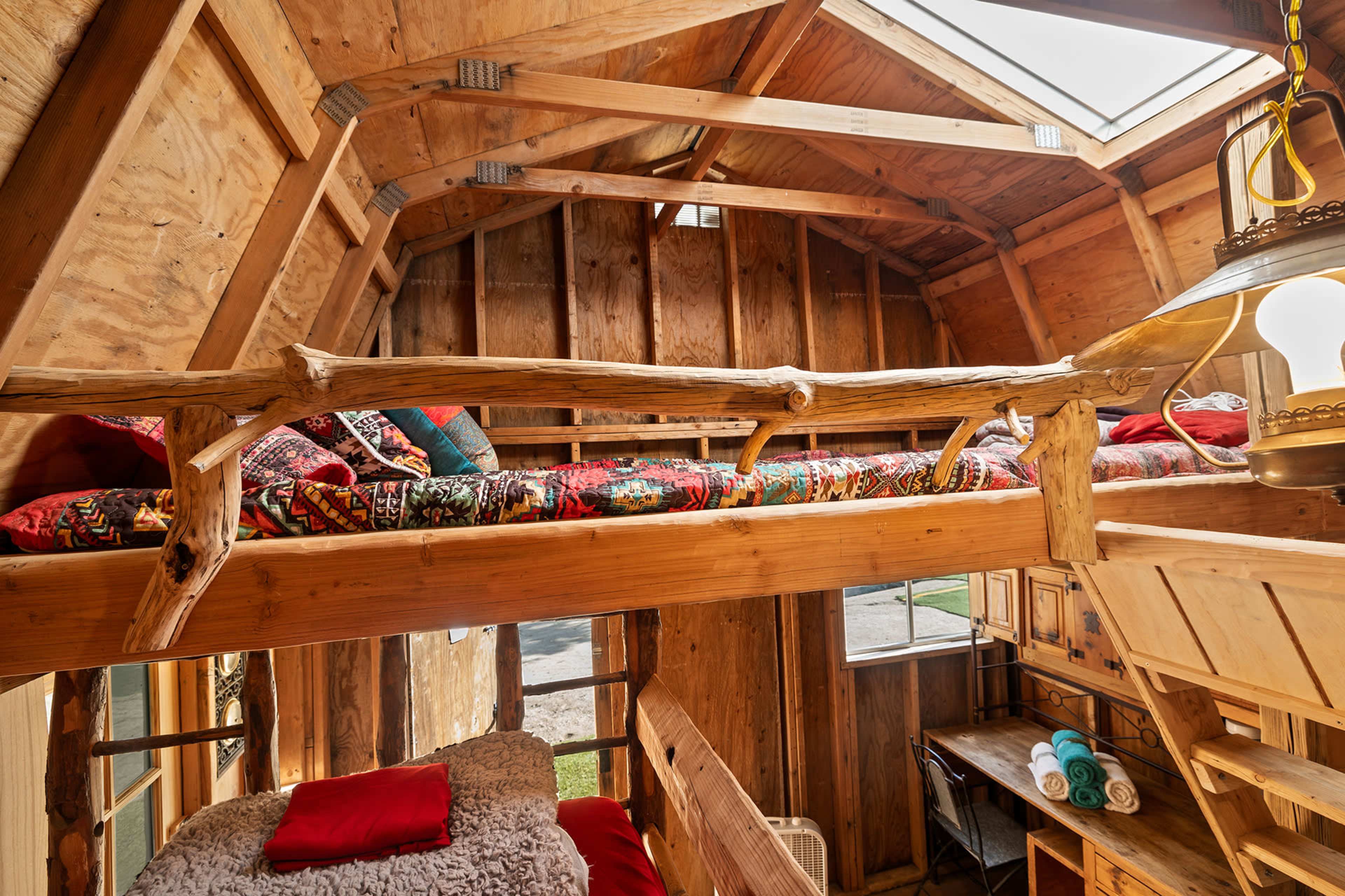 The image shows the interior of a rustic wooden cabin featuring a lofted sleeping area with colorful bedding and a cozy ground level with a plush bed and a small table.