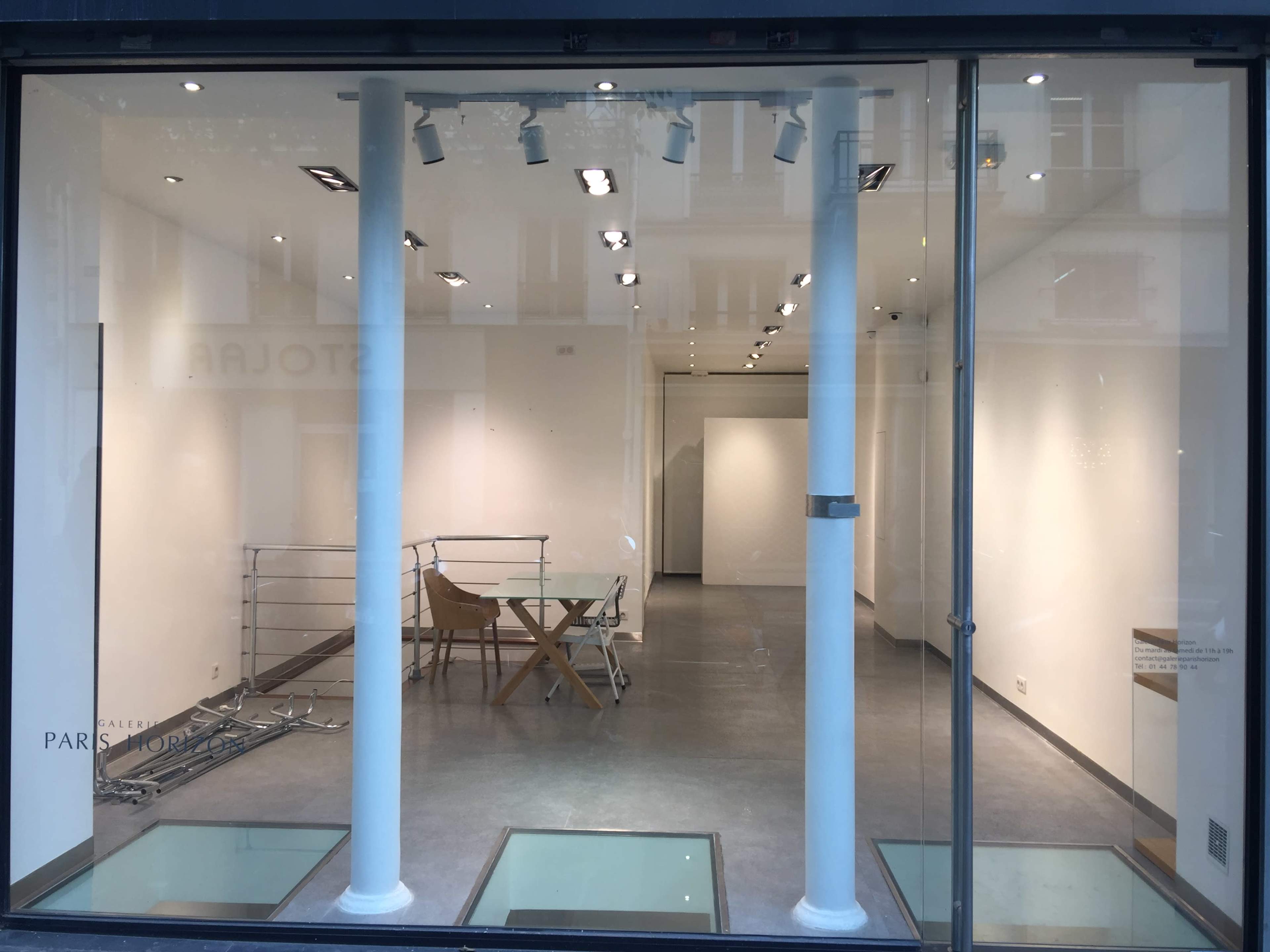 The image shows a minimalist, empty art gallery with white walls, polished floors, and a single table and chairs in the center.