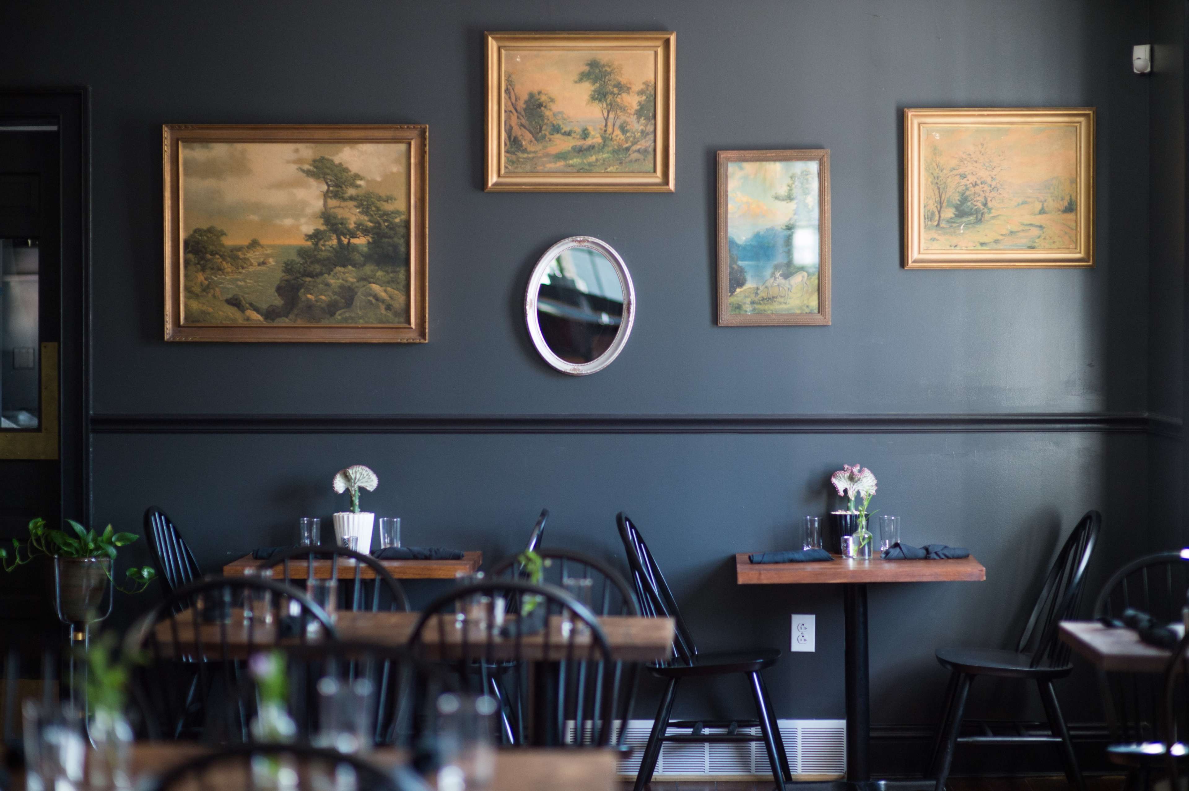 A dimly lit restaurant features dark walls adorned with framed landscape paintings and a circular mirror, with wooden tables and black chairs arranged for dining.