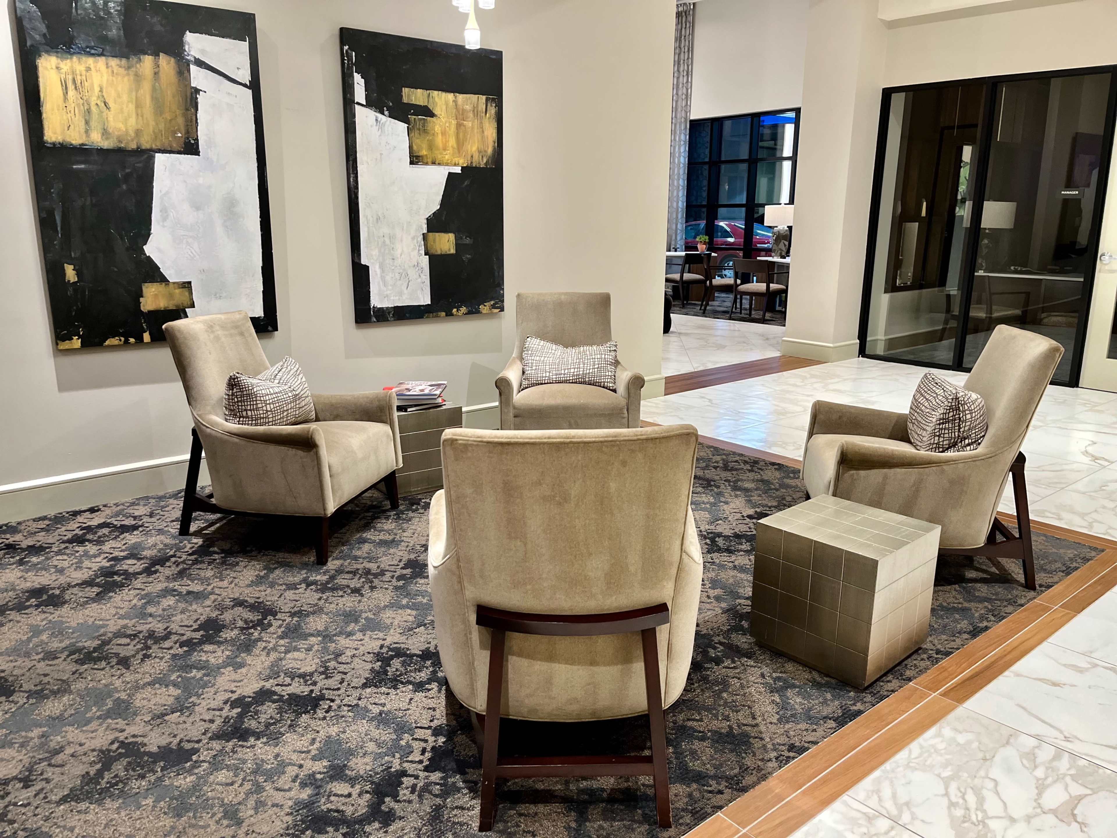 A modern seating area featuring four beige armchairs arranged around a small table on a patterned rug, with two abstract paintings hanging on the wall behind them.