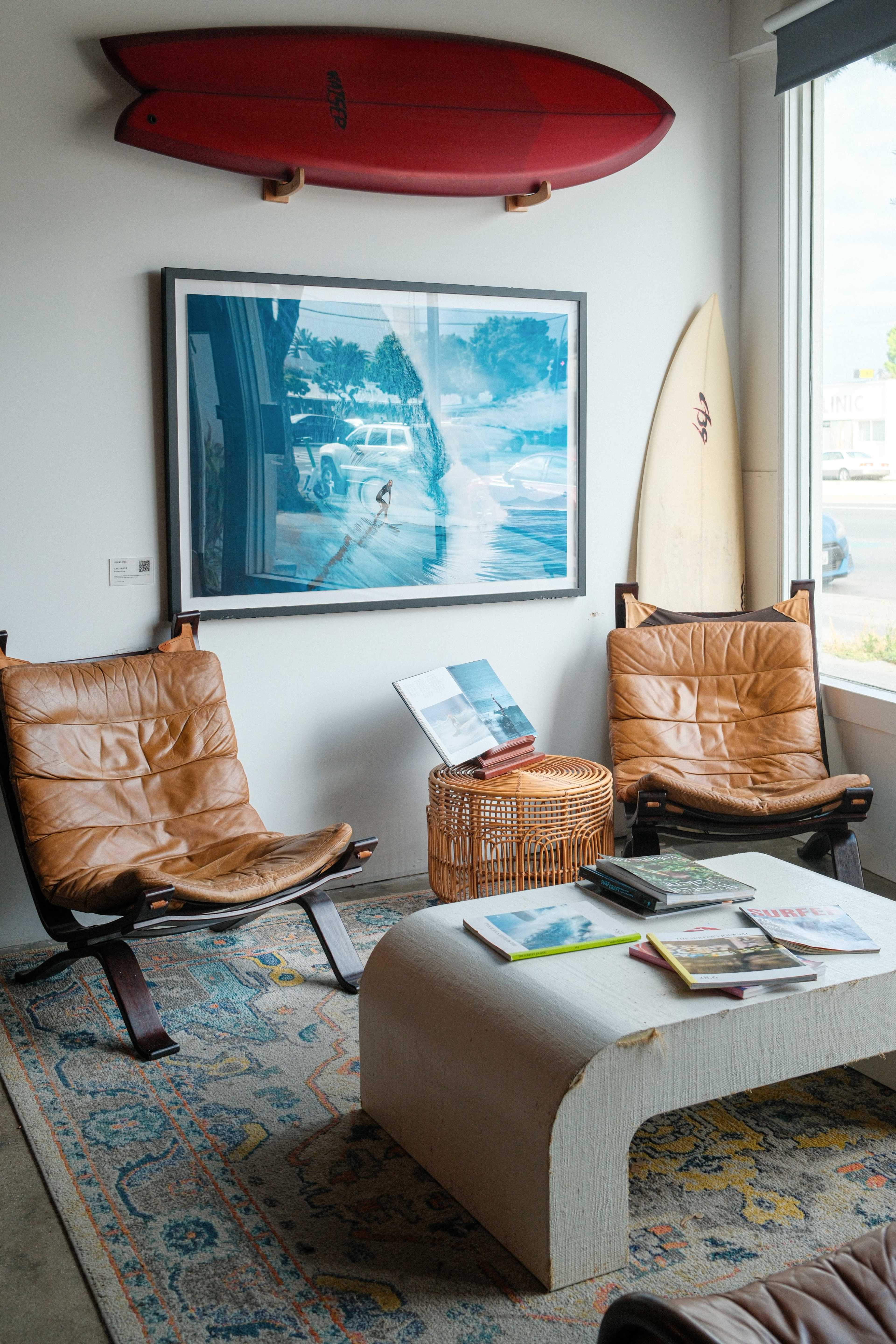 The image shows a cozy corner featuring two leather chairs, a coffee table with books, a woven basket, and surfboards mounted on the wall.