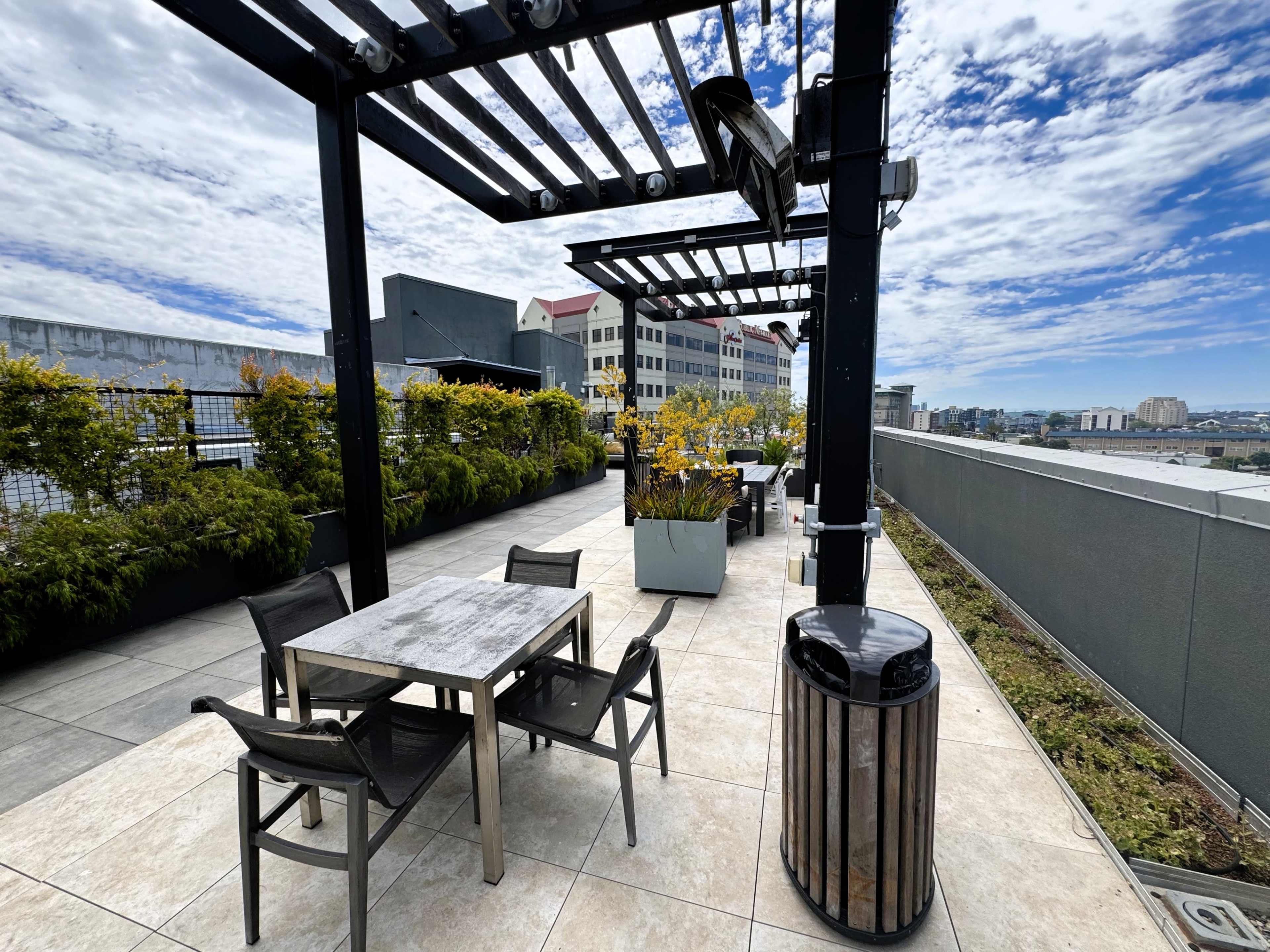 The image shows a rooftop terrace with patio furniture, planters, and a trash bin, under a partly cloudy sky.