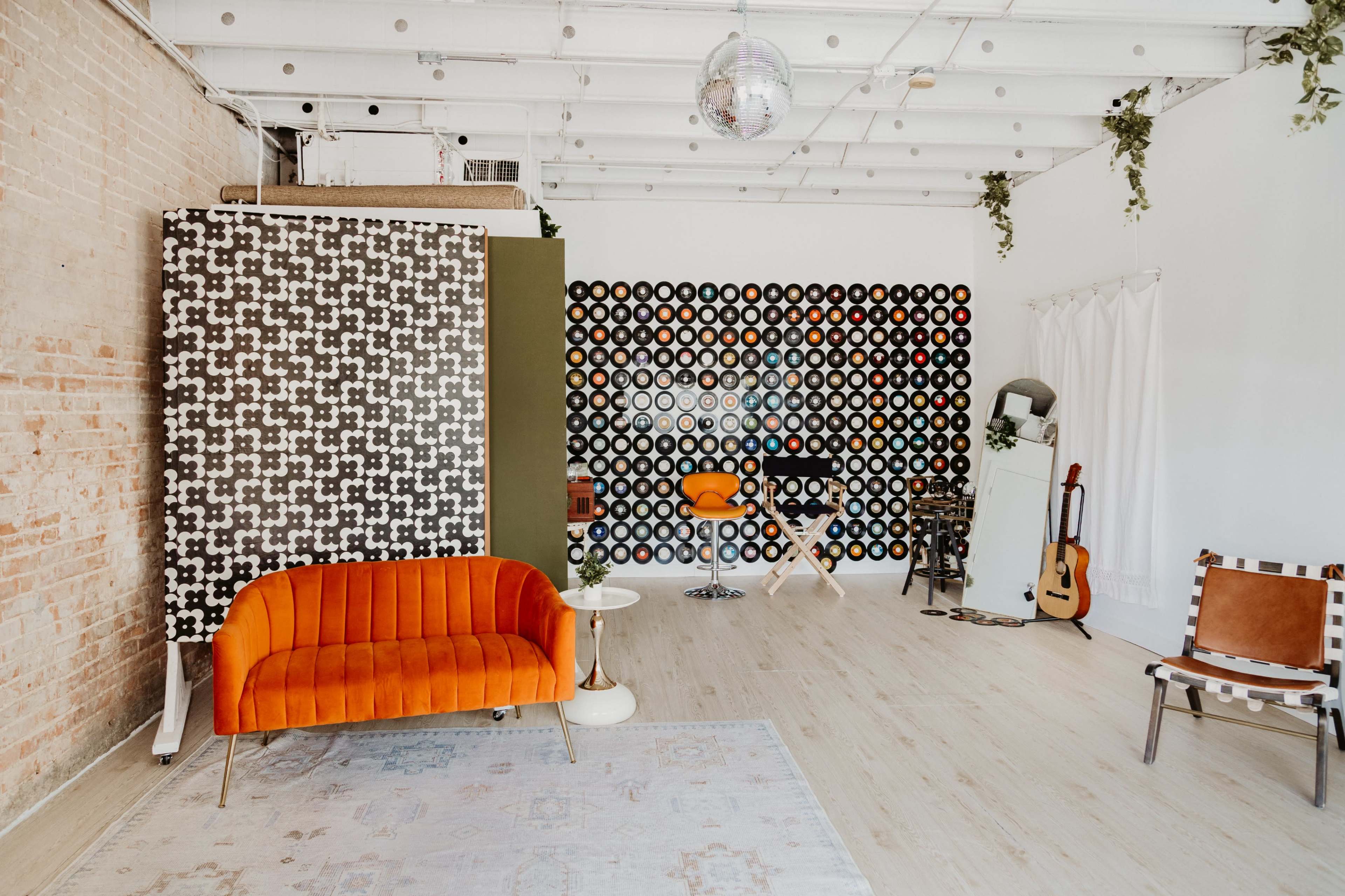 The image shows a spacious interior featuring an orange velvet couch, a patterned wall with colorful circles, a small table, and musical instruments in a well-lit studio space.