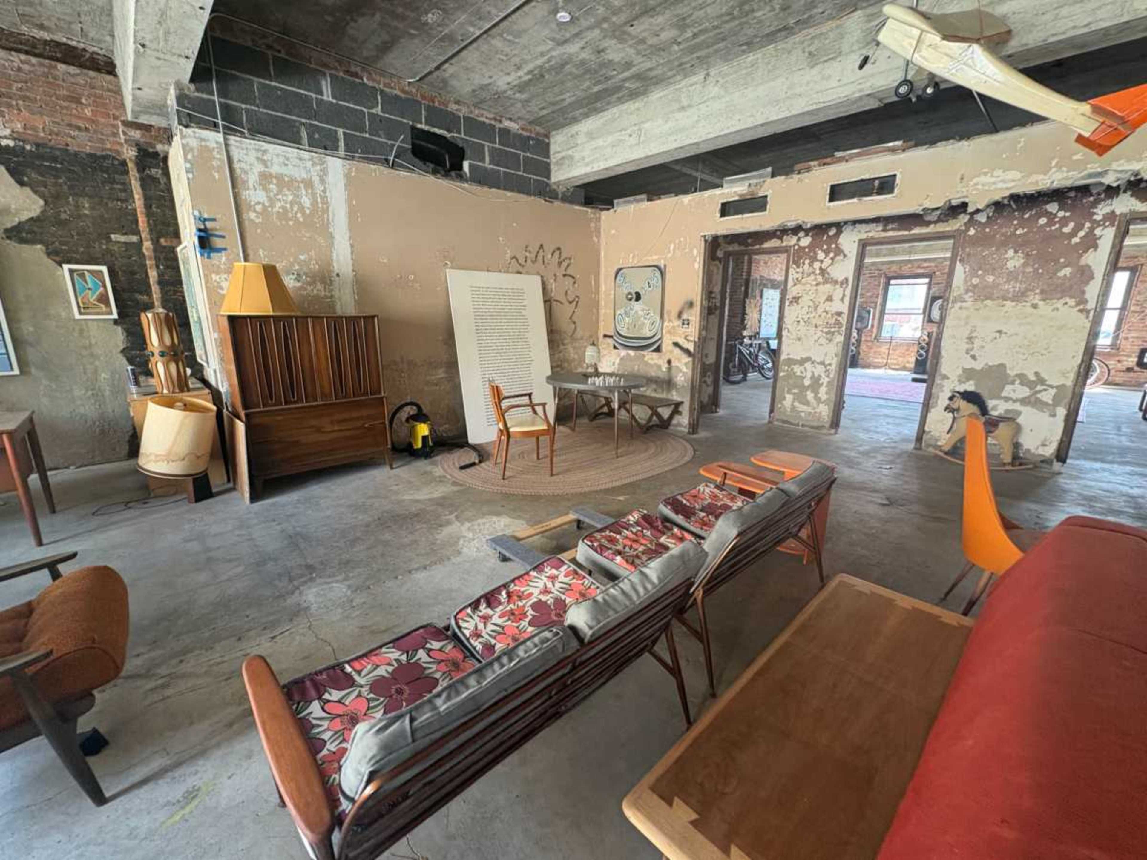 The image depicts a spacious, unfinished room with exposed brick walls, various pieces of mid-century modern furniture, and a distinct vintage aesthetic.