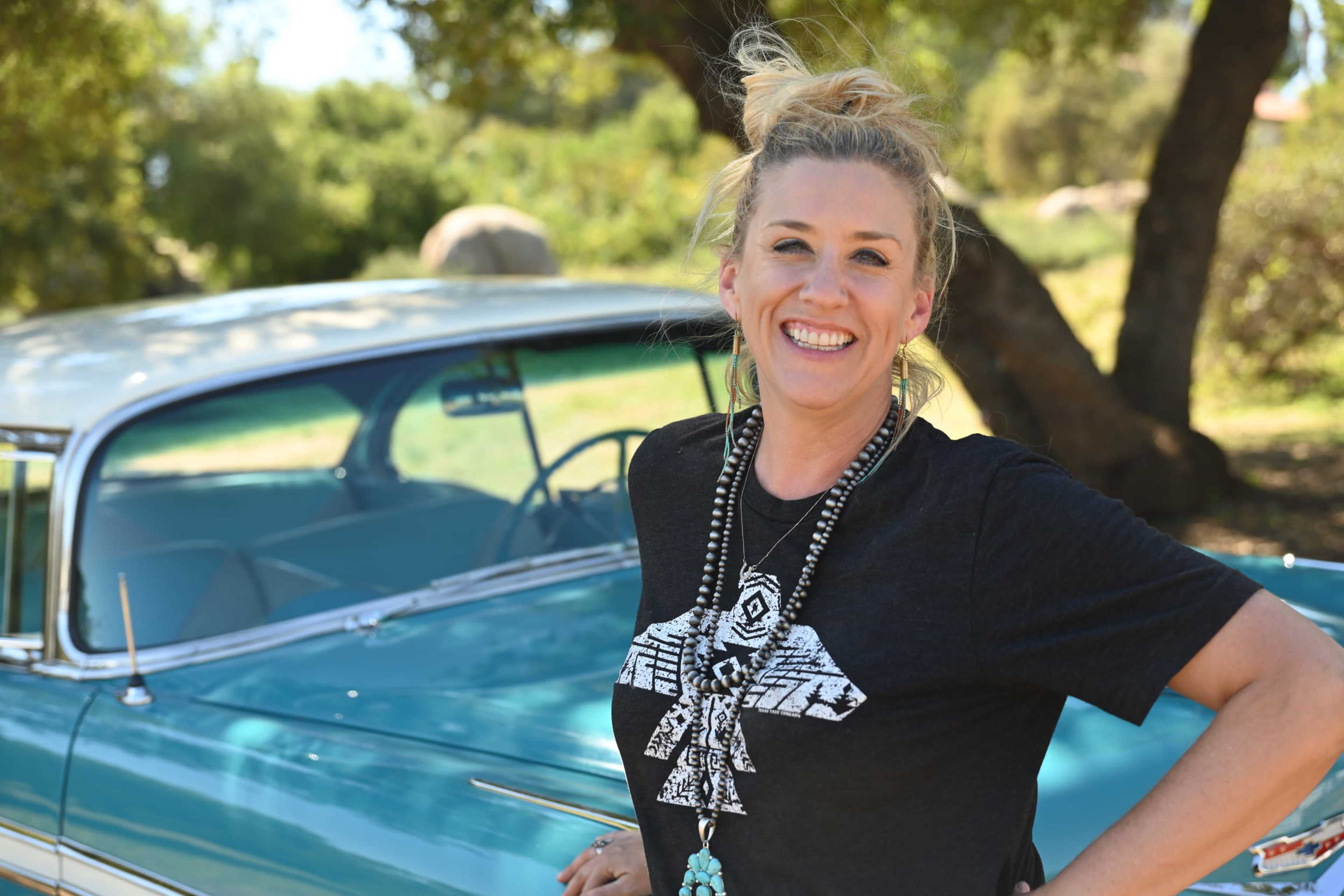 A woman stands smiling beside a vintage blue car in a natural outdoor setting.