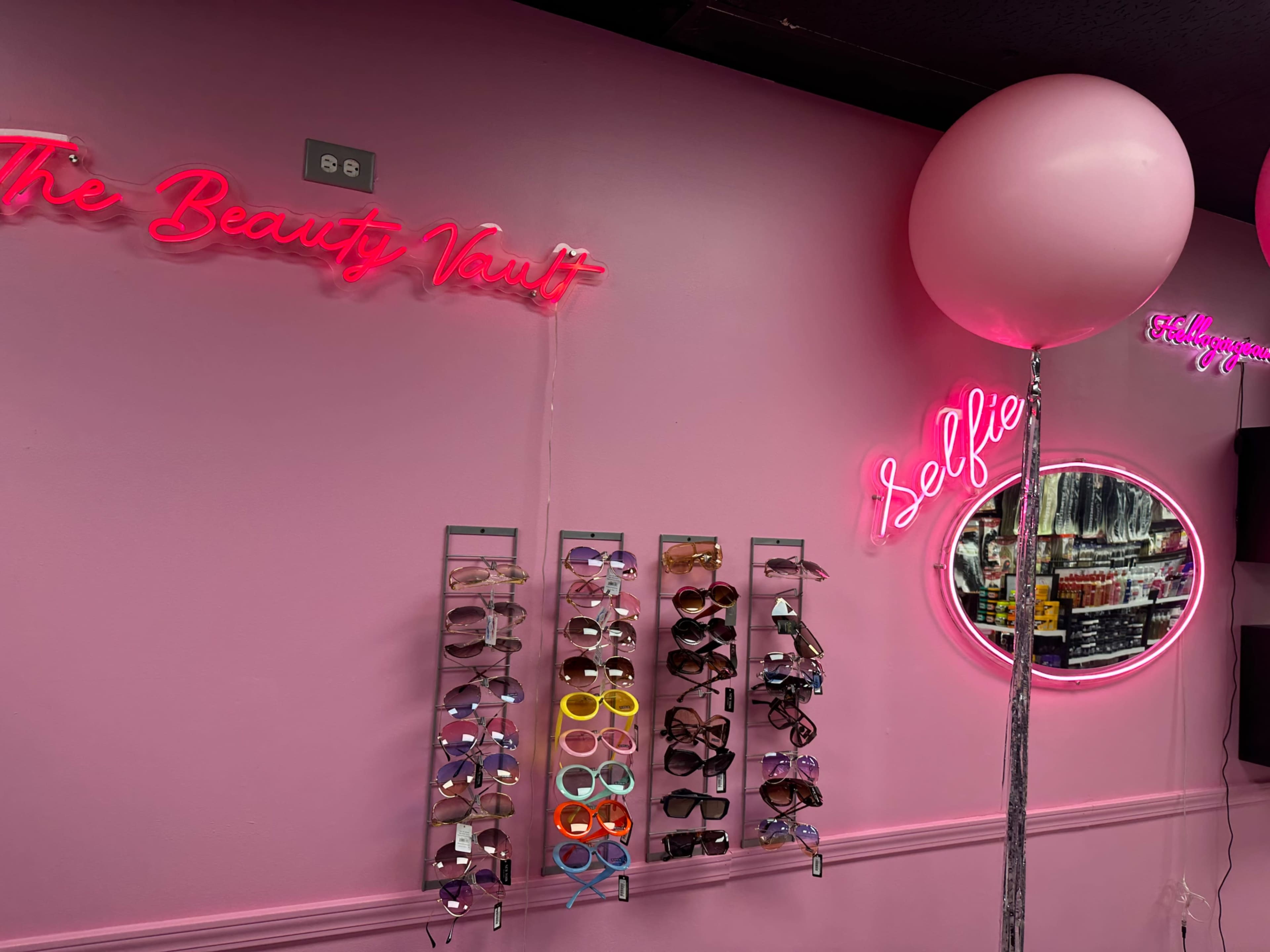 A brightly colored pink wall with neon signs, a circular mirror, and a display rack of sunglasses.