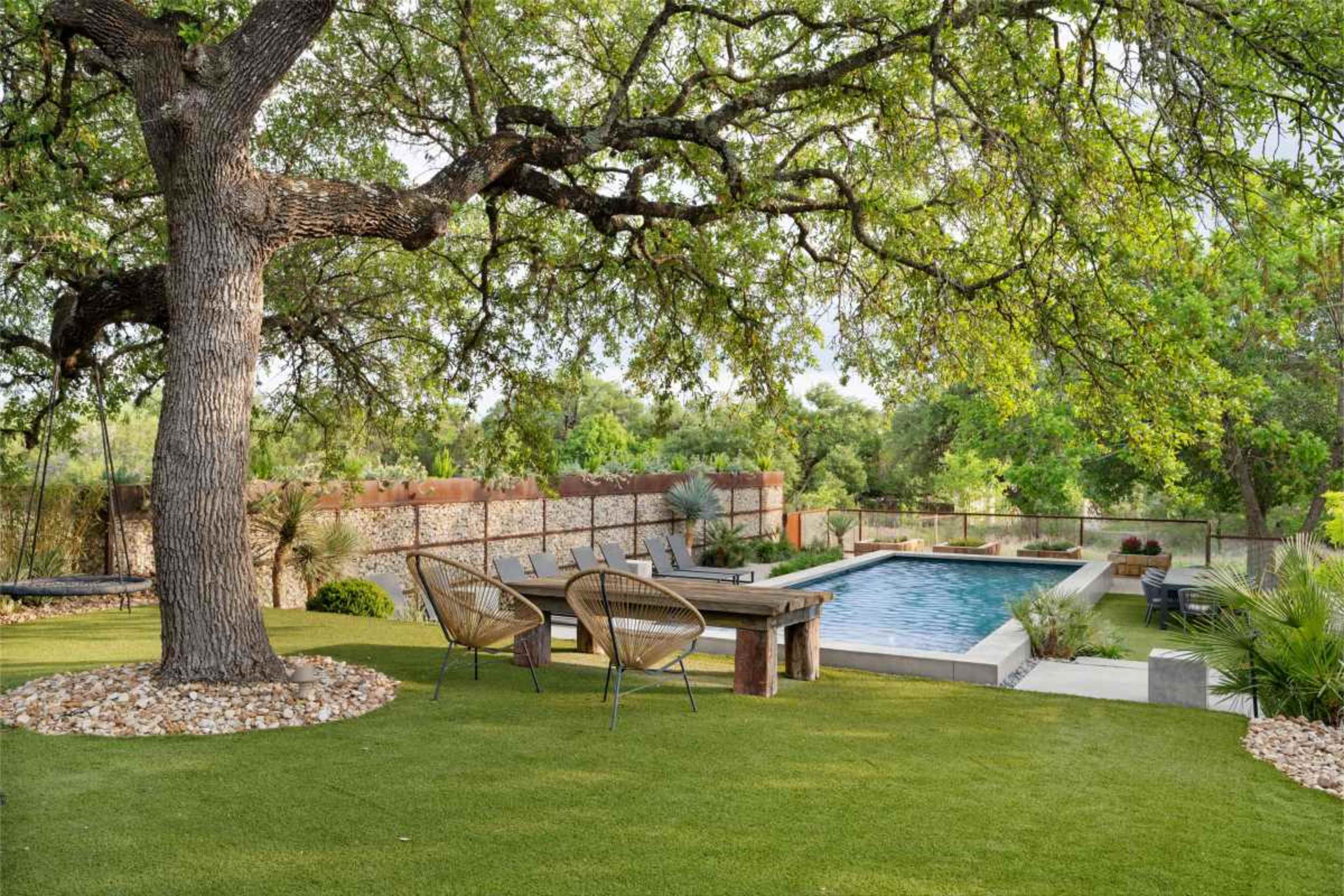 The image depicts a backyard scene featuring a swimming pool, a wooden dining table with two chairs, and an expansive tree providing shade in a landscaped area.