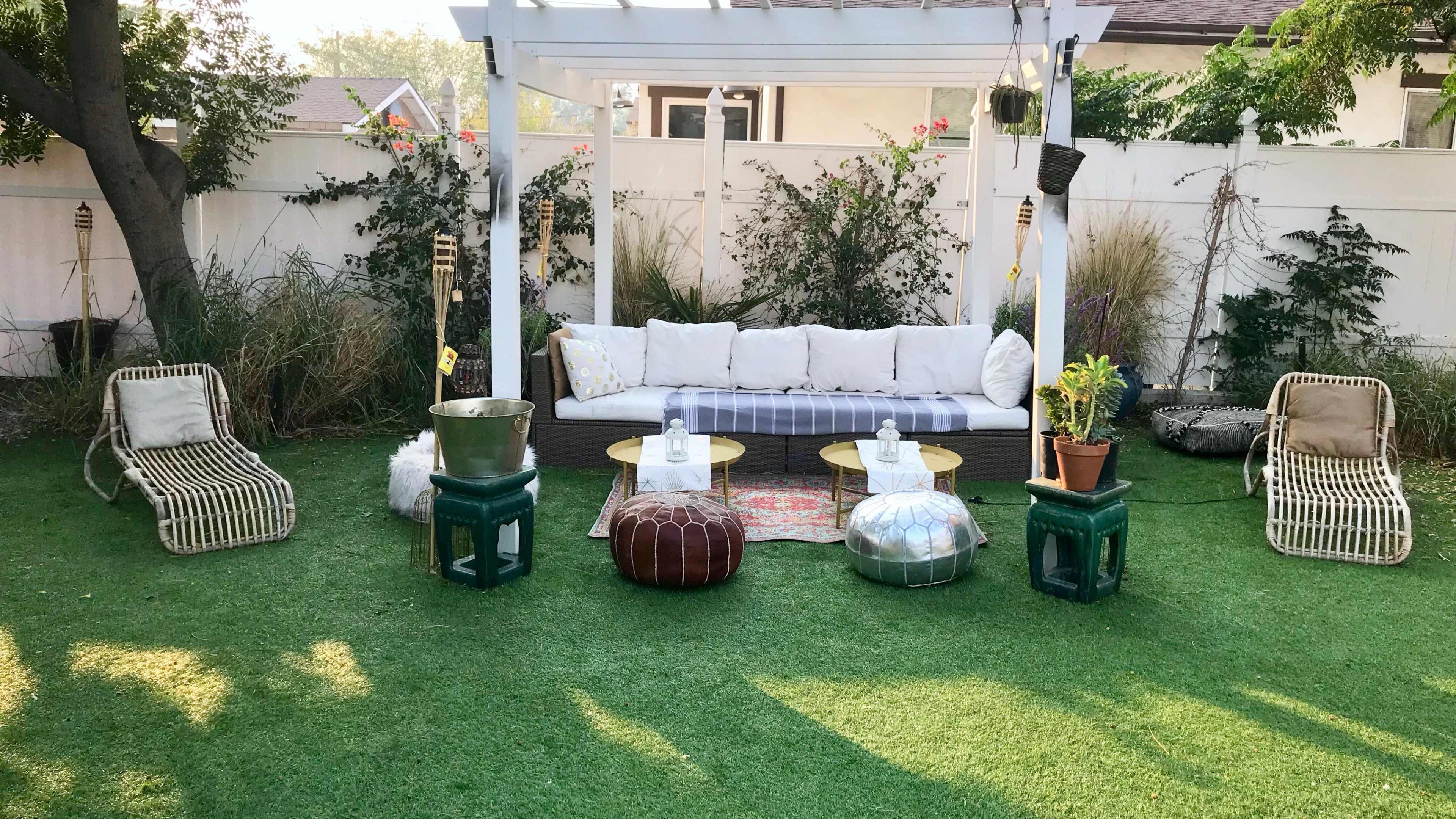 A backyard seating area with a white sofa, round tables, and decorative poufs, surrounded by green grass and plants.