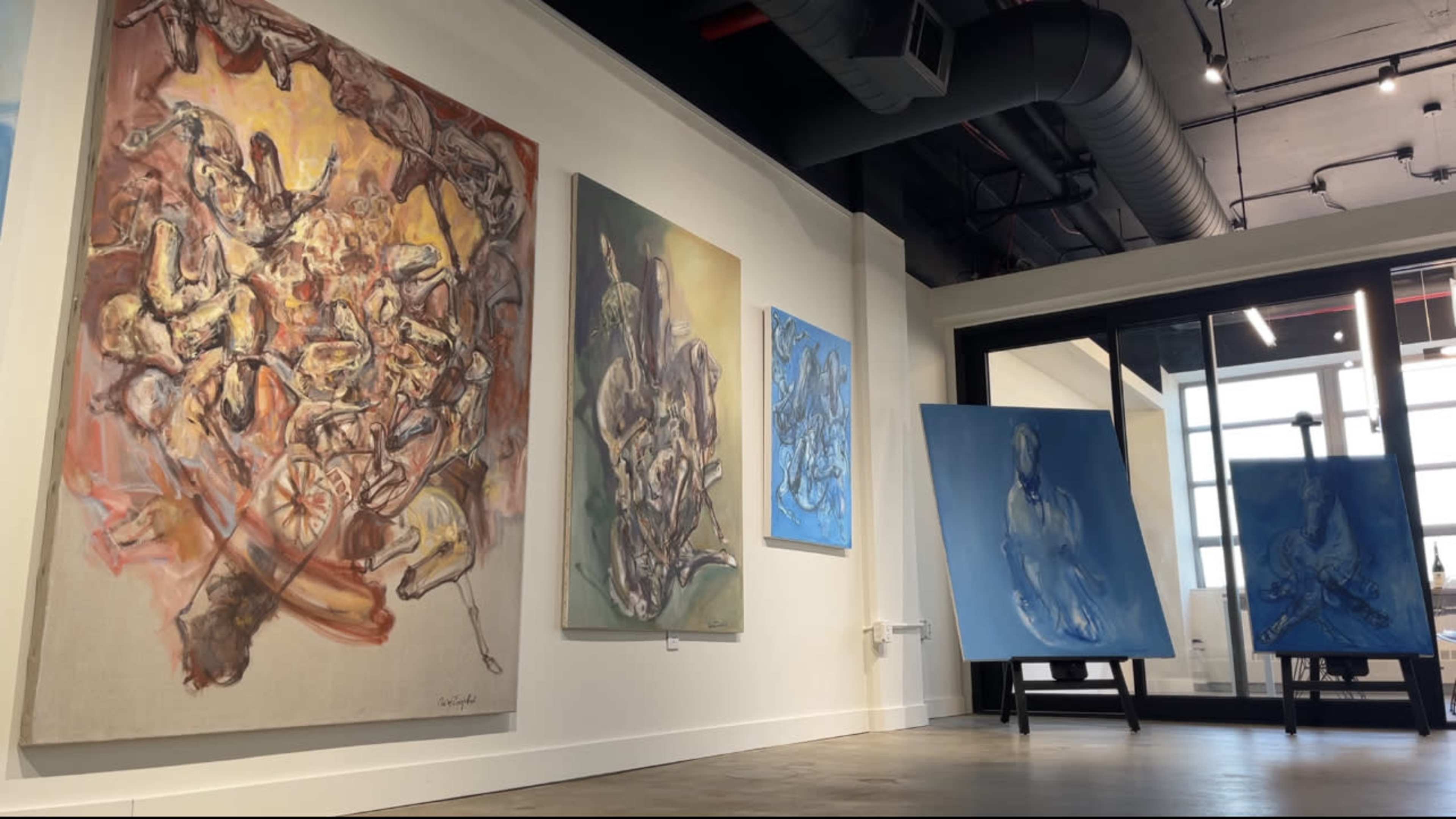 An art gallery featuring several large paintings on the walls, including abstract and figurative works in various colors.