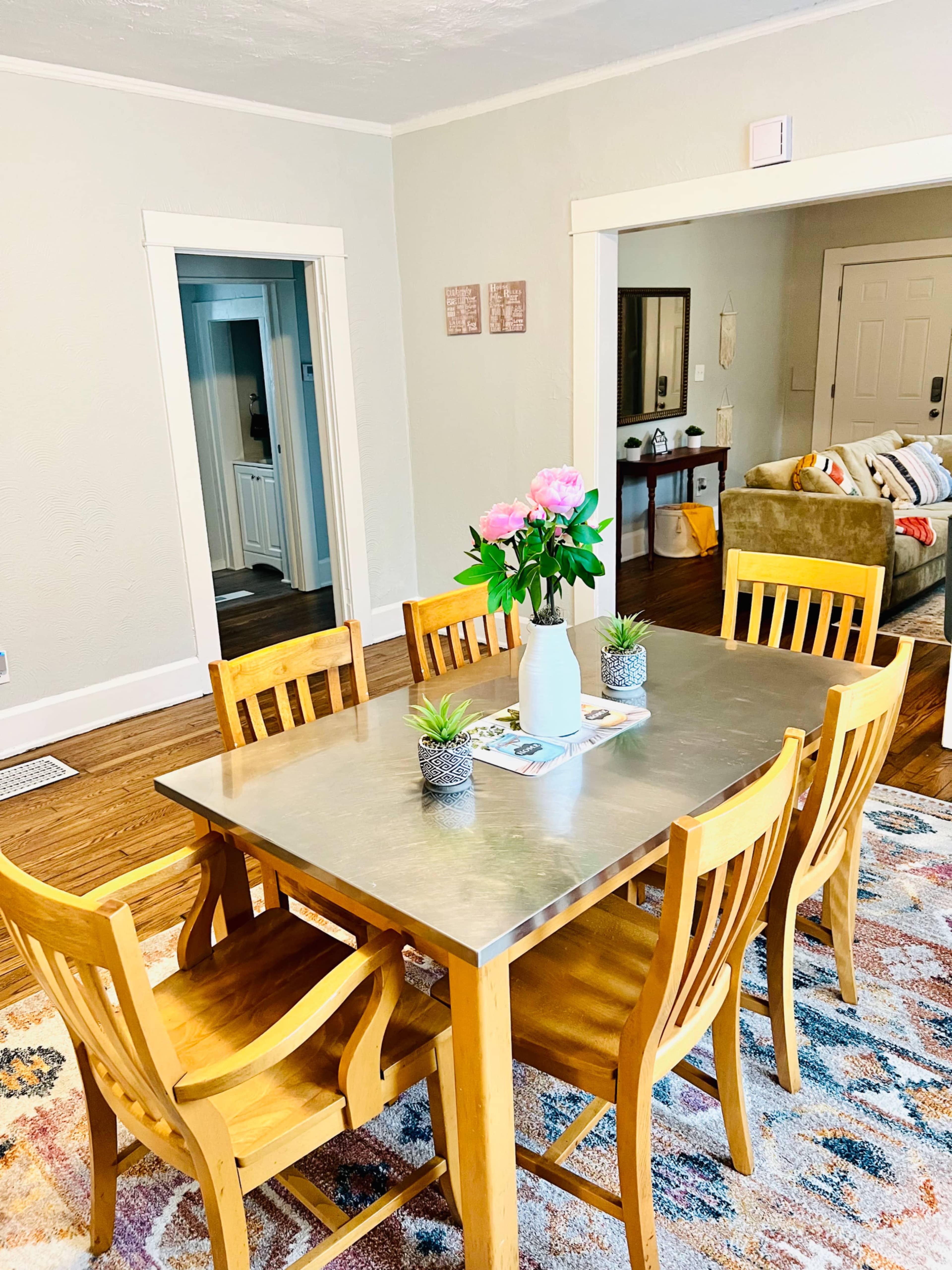 A modern dining room features a metal table surrounded by wooden chairs, with a vase of flowers and small plants on the table, and a cozy living area visible in the background.