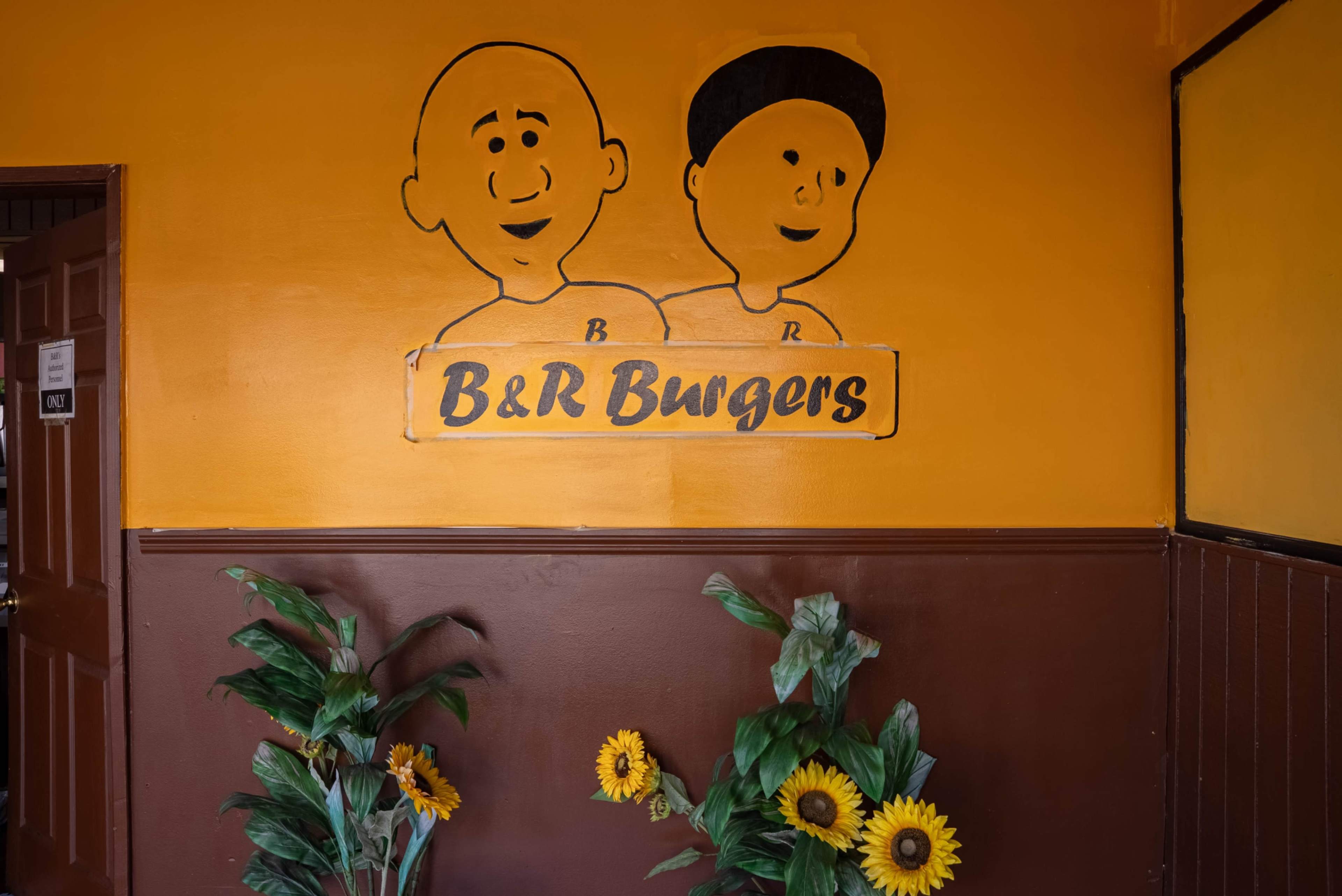 The image shows a brightly colored wall featuring a mural of two cartoon characters alongside a sign that reads "B&R Burgers," with potted sunflowers placed at the base.