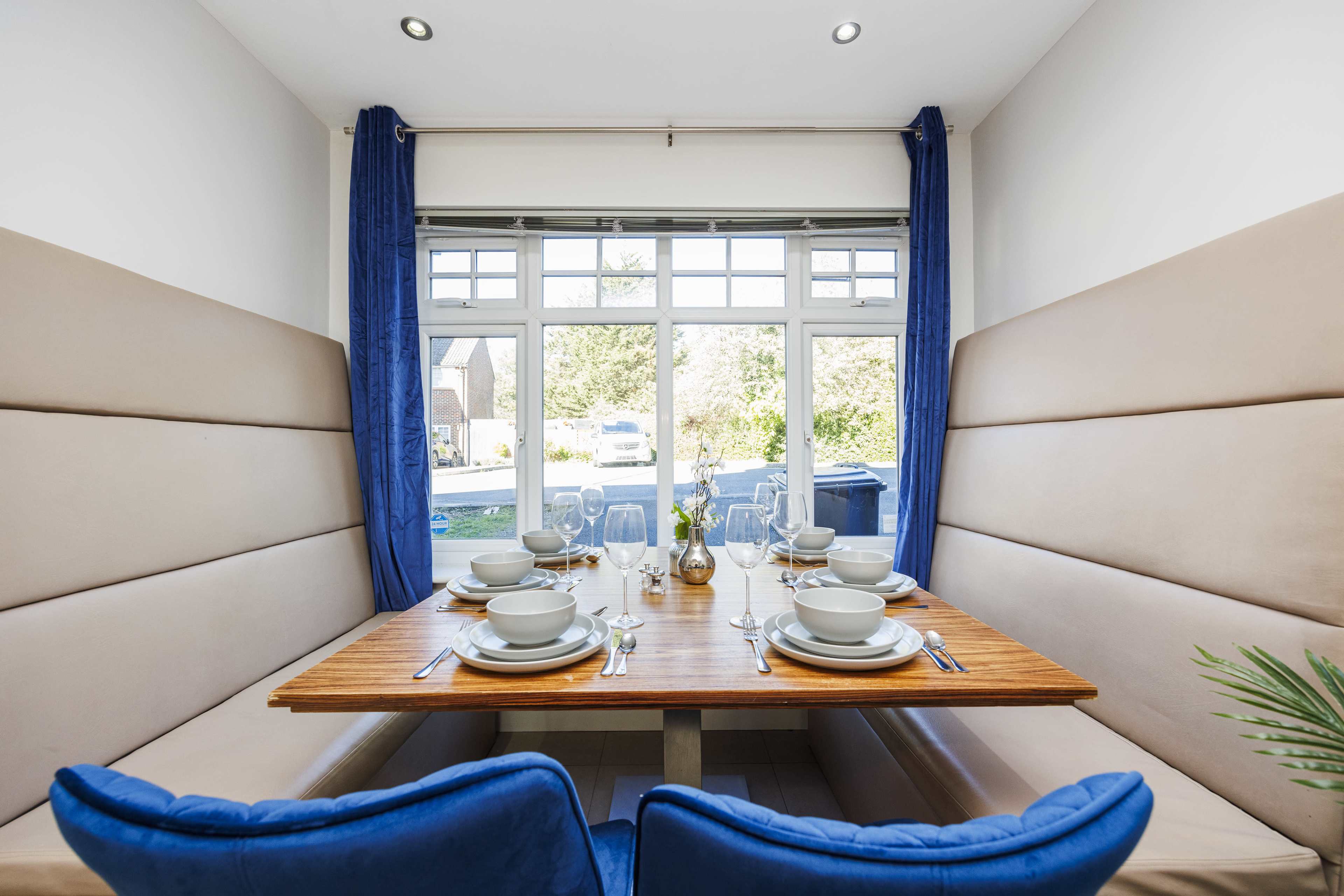 A dining area featuring a wooden table set with dishes and cutlery, flanked by upholstered seating and large windows allowing natural light.