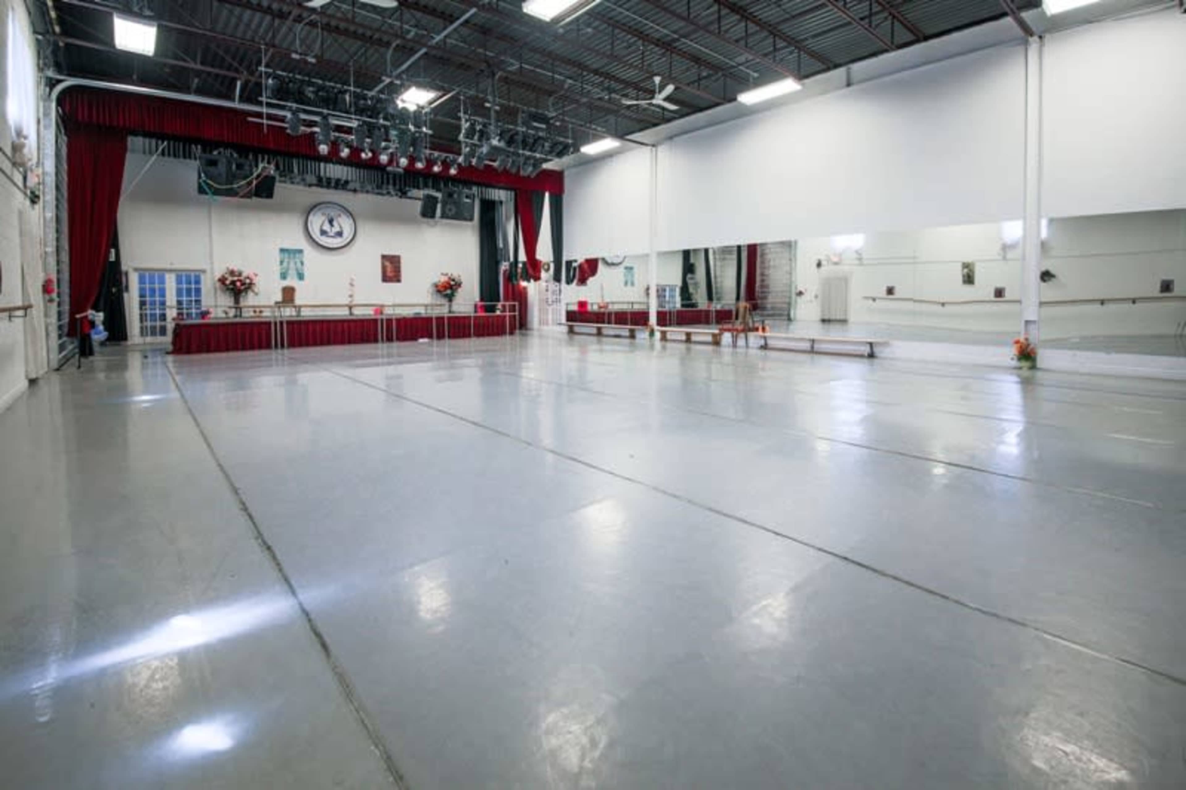 A spacious dance studio with a wooden barre, mirrored walls, and a stage area at one end.