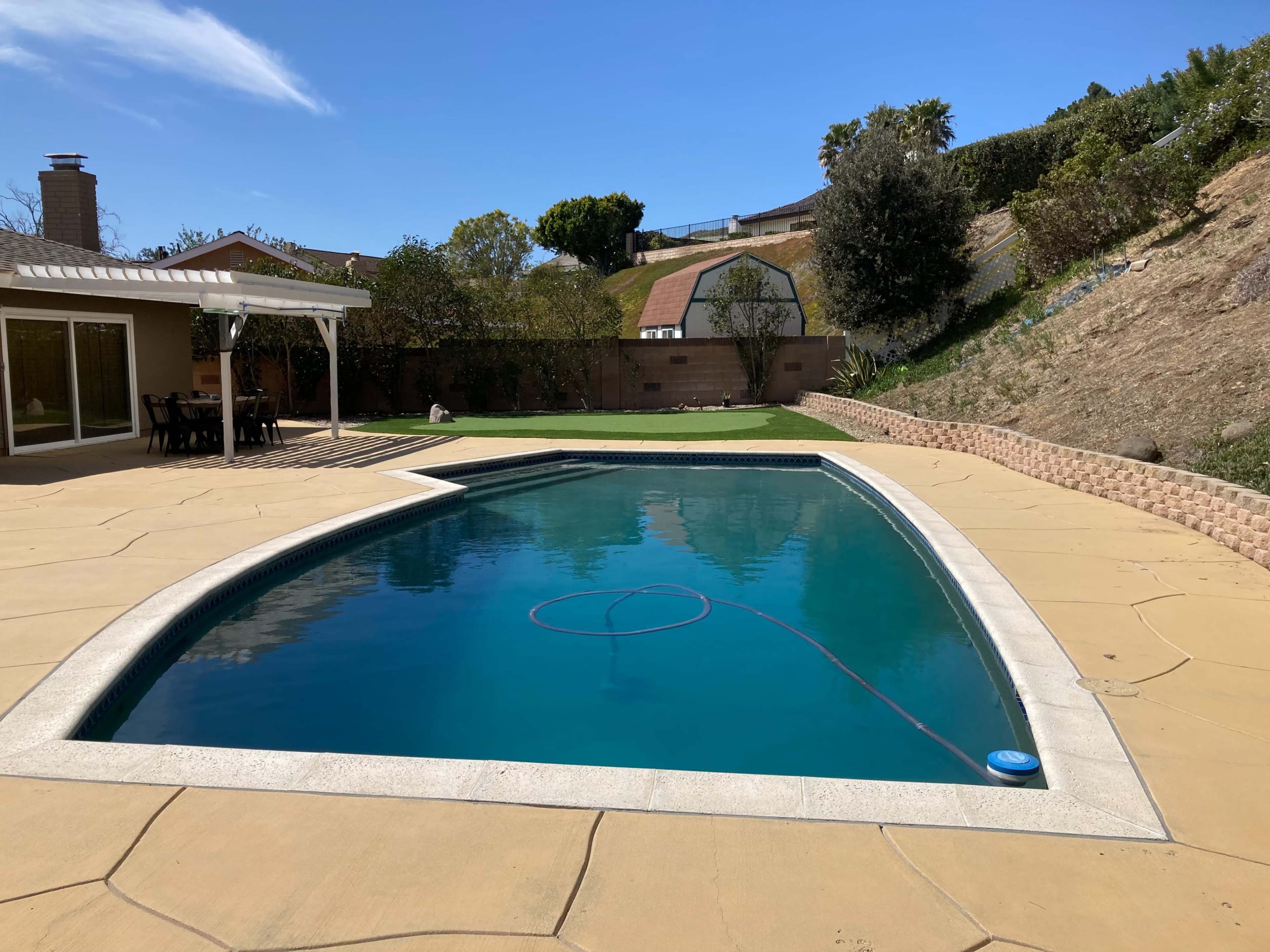 A rectangular swimming pool surrounded by a concrete deck, with a green lawn and hillside in the background.