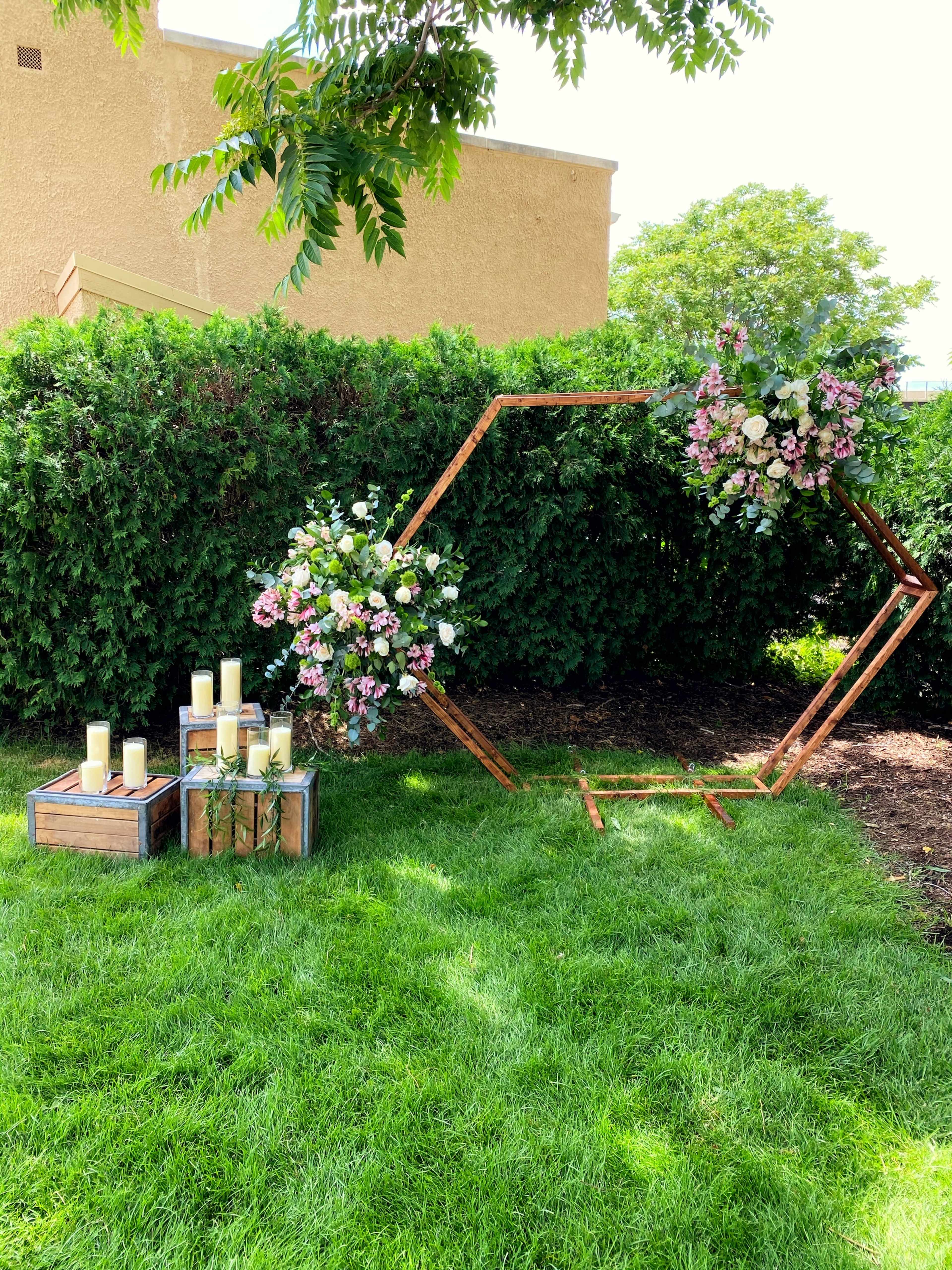 The image shows a hexagonal wooden frame adorned with colorful flowers, positioned near a cluster of candles on wooden crates, set against a backdrop of dense greenery.