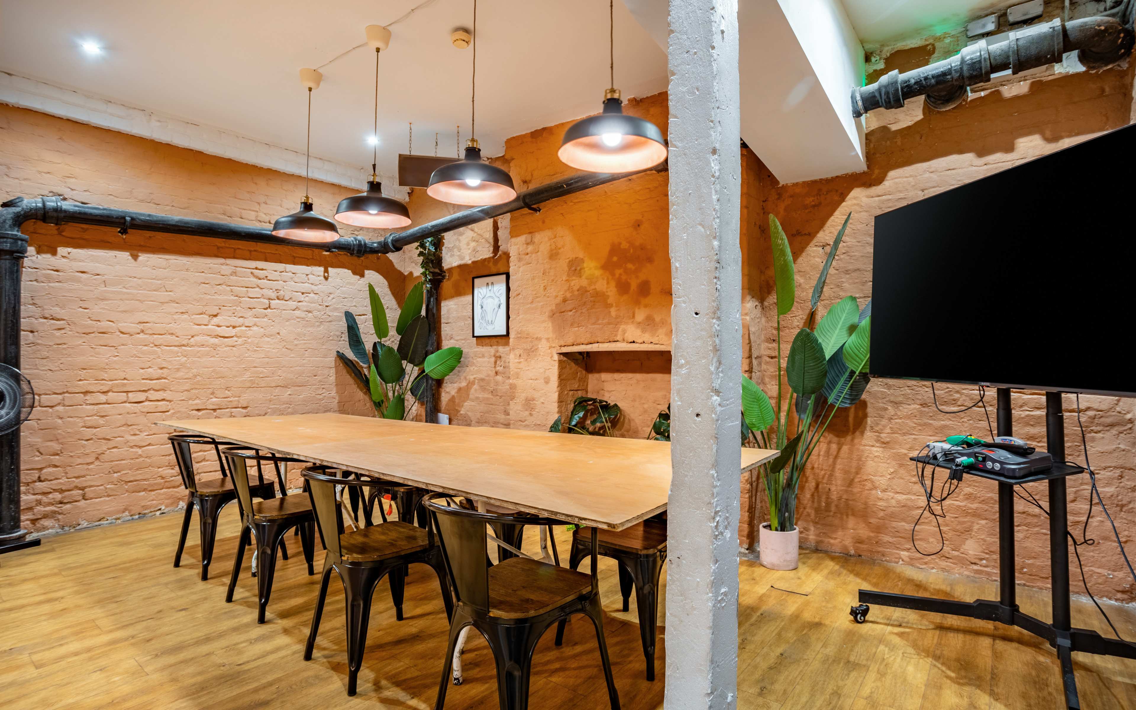 A long wooden table surrounded by black metal chairs is illuminated by hanging lights in a room with an orange brick wall and potted plants.