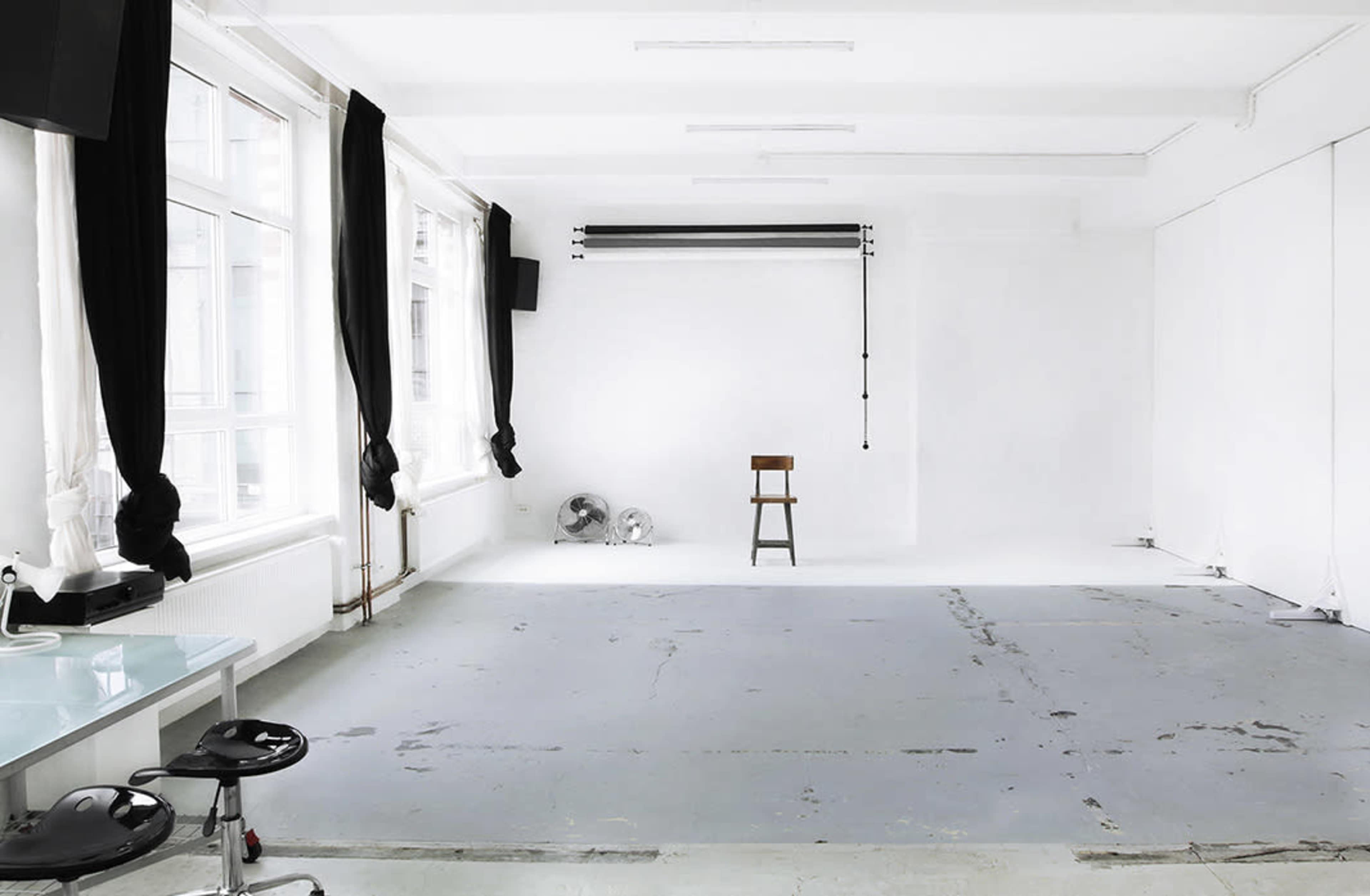 A spacious, minimalist room with white walls, large windows, a single wooden chair, and a few scattered fans.