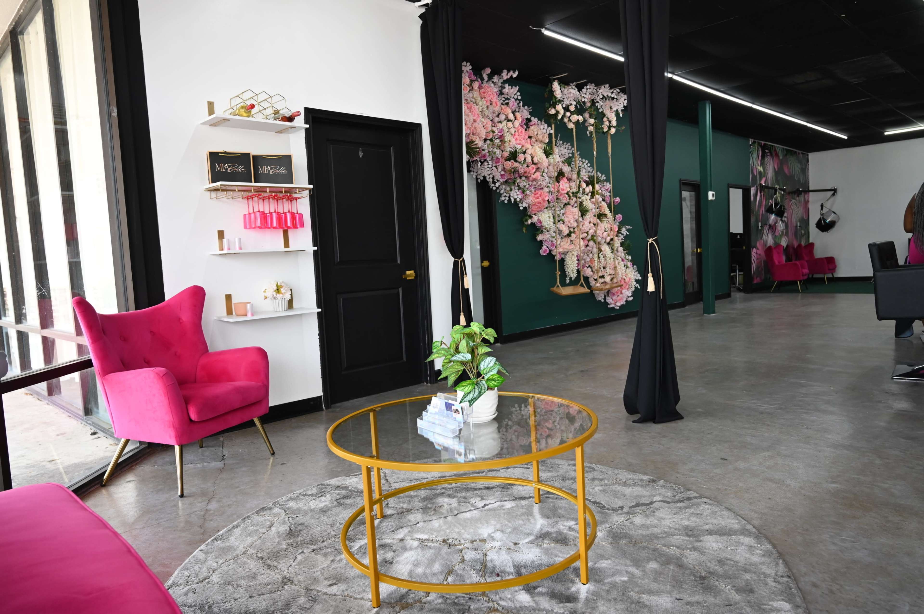 The interior of a salon features a pink chair, a glass coffee table on a circular rug, and a wall adorned with floral decorations.