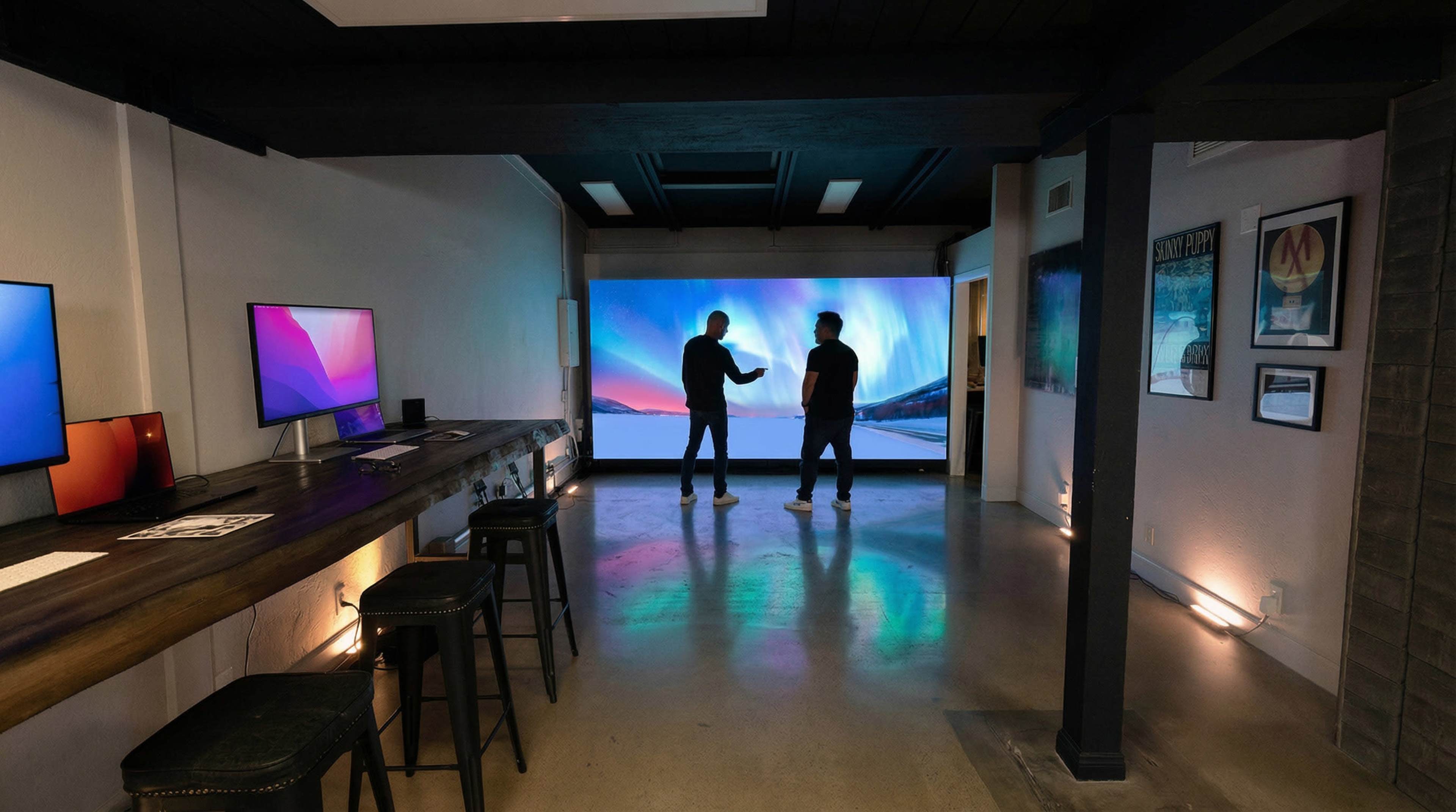 Two individuals stand facing each other in a modern room with a large projection screen displaying colorful graphics and a bar-style table with computers on one side.