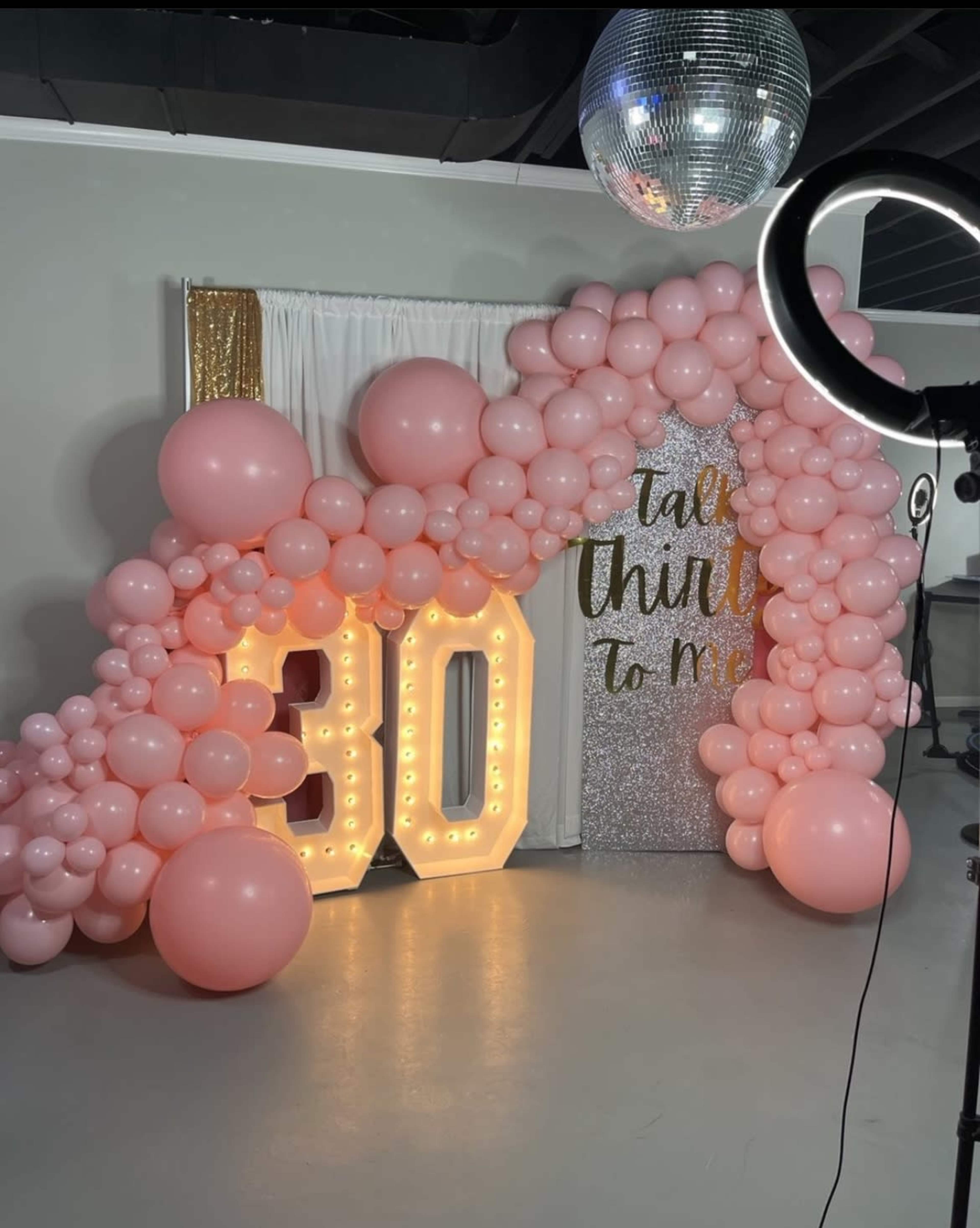 A festive backdrop featuring large illuminated numbers "30" surrounded by pink balloons and a glittery panel with the text "Talk thirty to me."