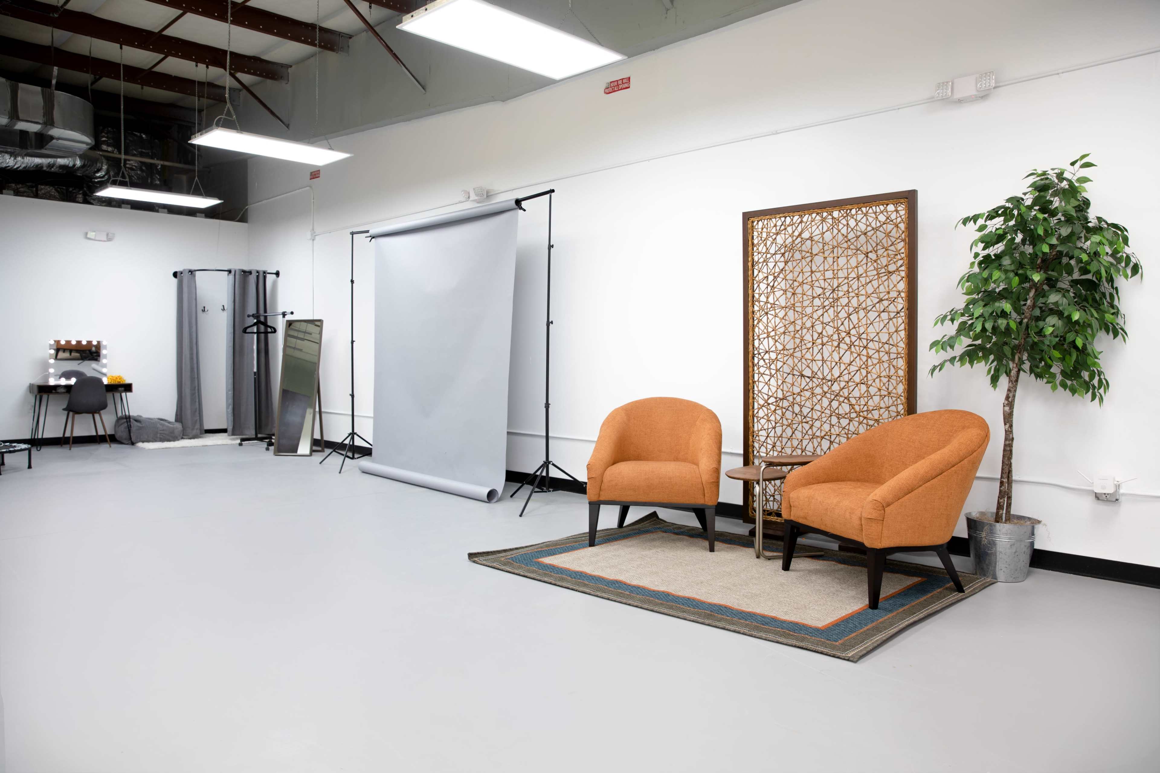 The scene shows a modern studio space featuring two orange chairs, a small table, a patterned rug, a backdrop stand with a gray backdrop, and a decorative screen, accompanied by a plant.