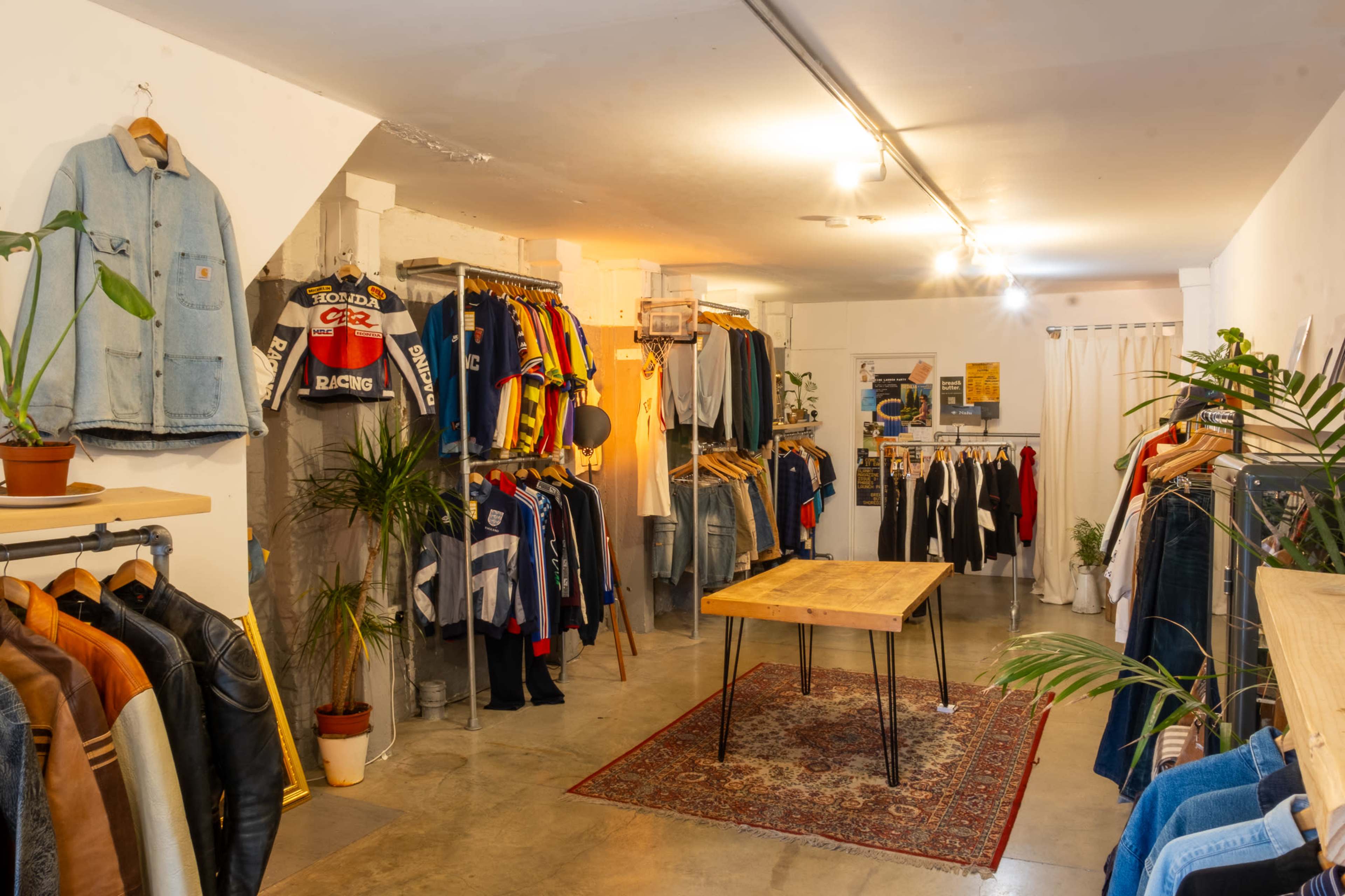 A vintage clothing store features racks of colorful apparel, neatly arranged along the walls, with a central wooden table surrounded by plants.