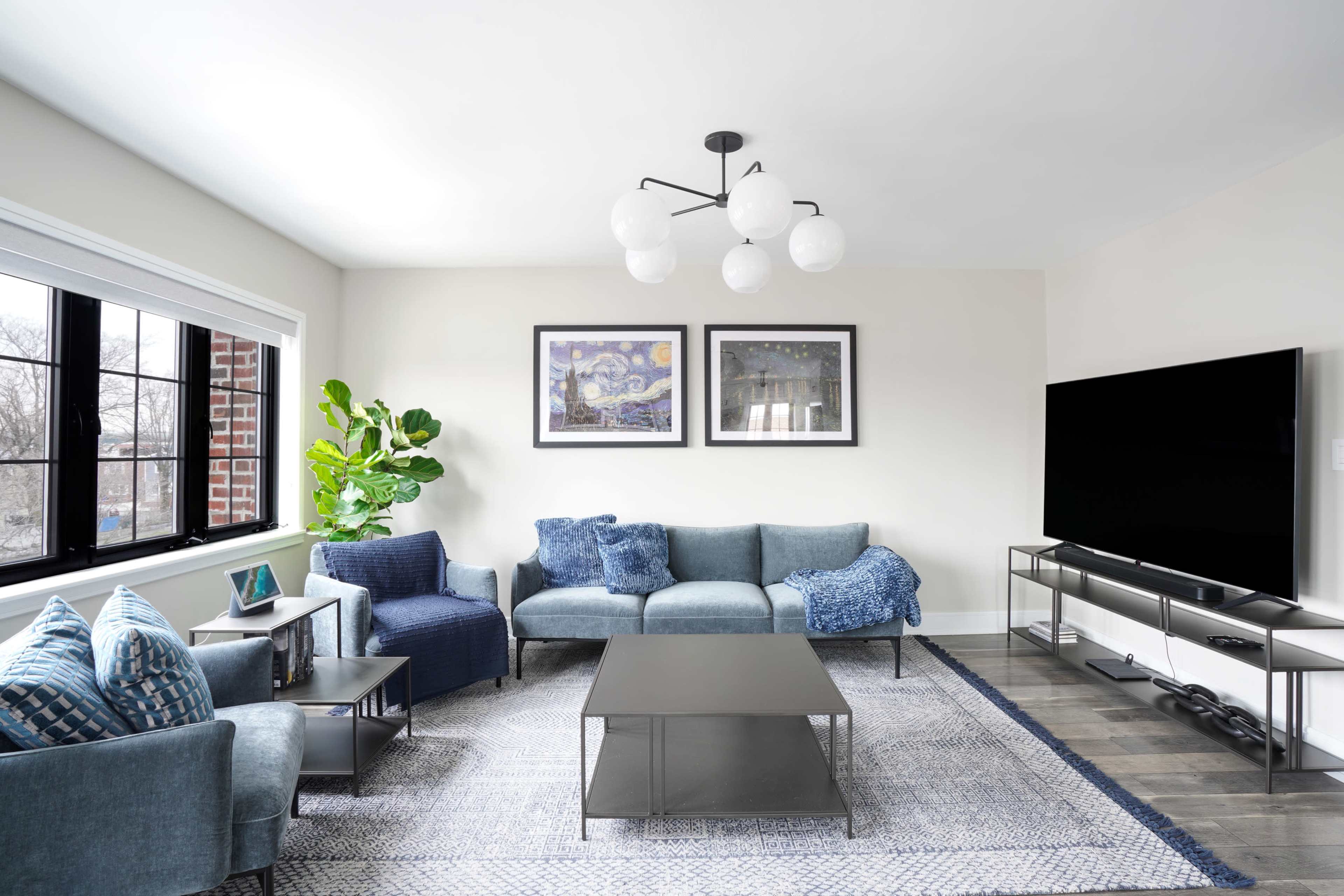 The living room features a sectional sofa, a coffee table, a large television on a stand, and two framed artworks on the wall, with a large window allowing natural light and a potted plant in the corner.