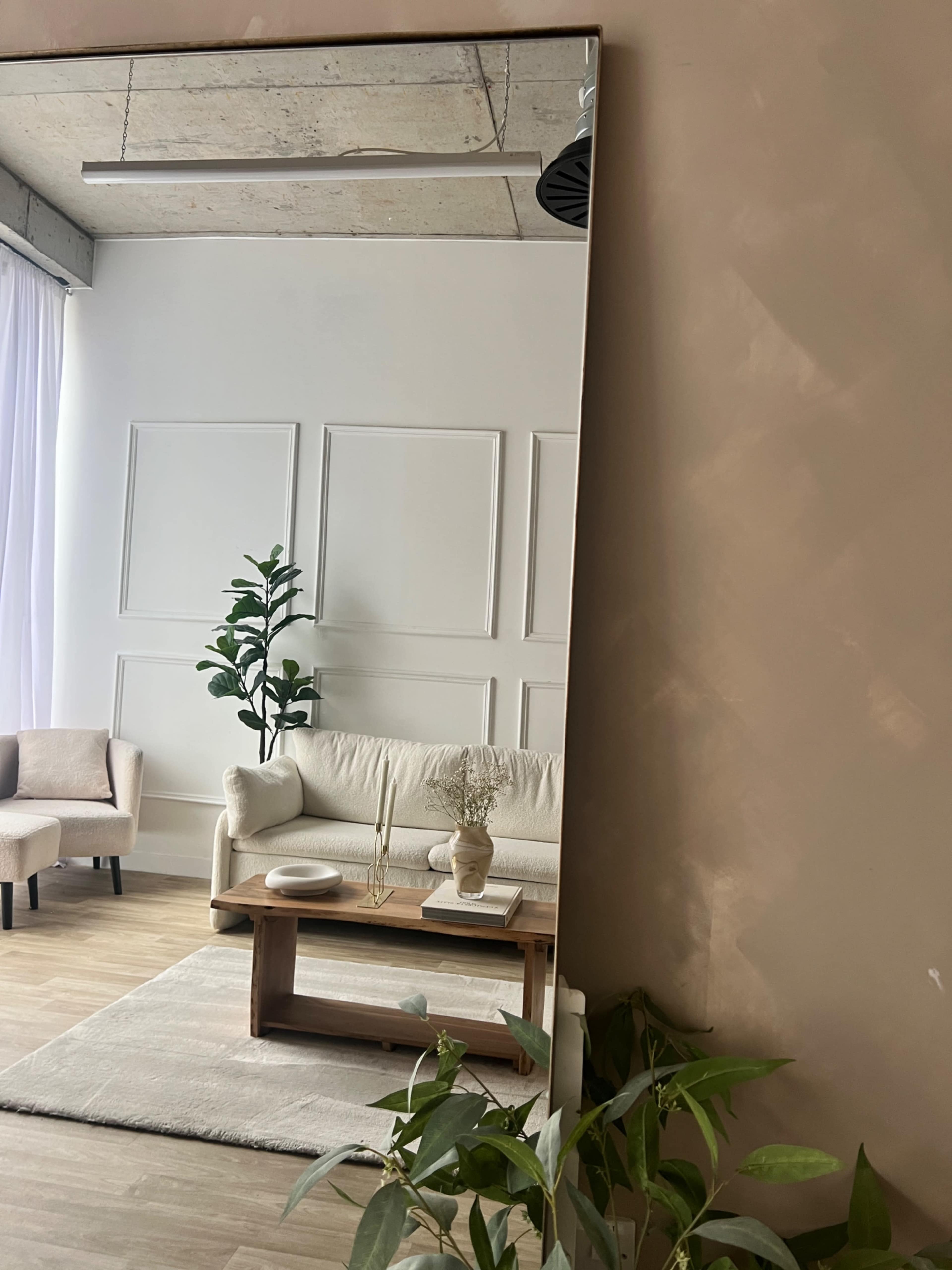 A modern living room with a white sofa, a wooden coffee table, and a decorative plant, reflected in a large mirror on the wall.