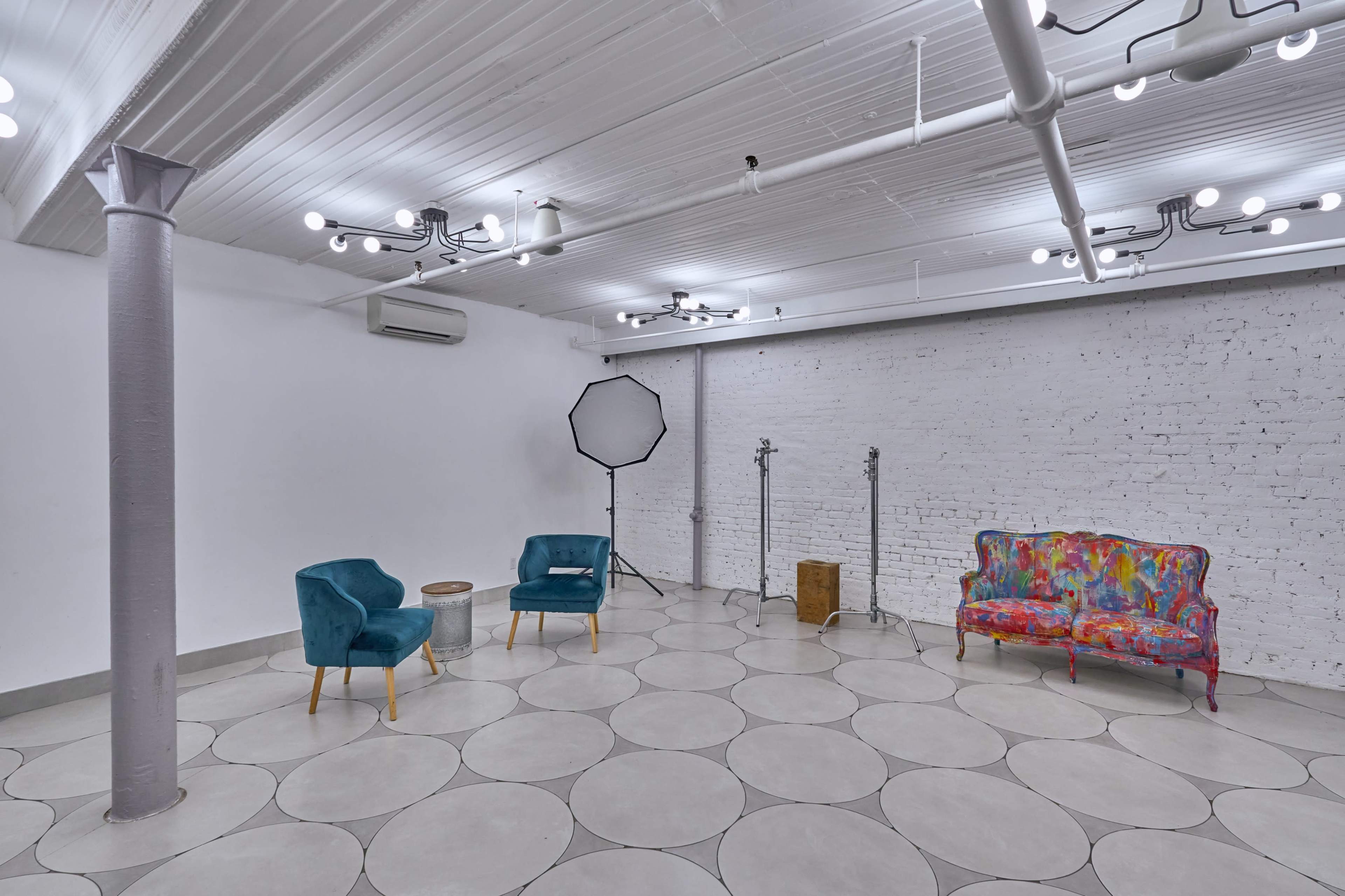 An empty studio space with a patterned floor, two teal chairs, a colorful sofa, and lighting equipment against a white brick wall.