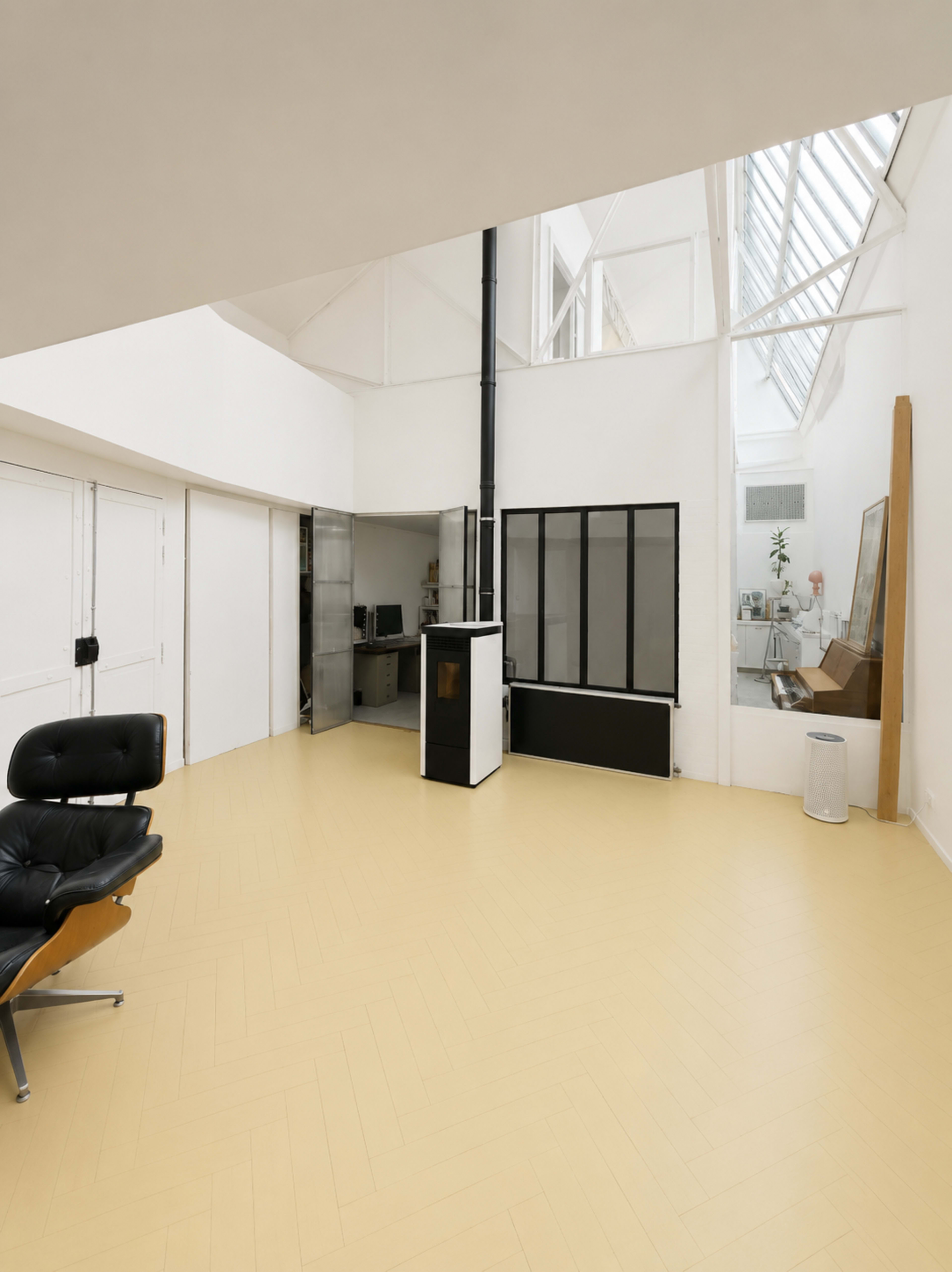 The image shows a minimalist interior space with a pale yellow floor, a black chair, and a partially visible office area with a large window.