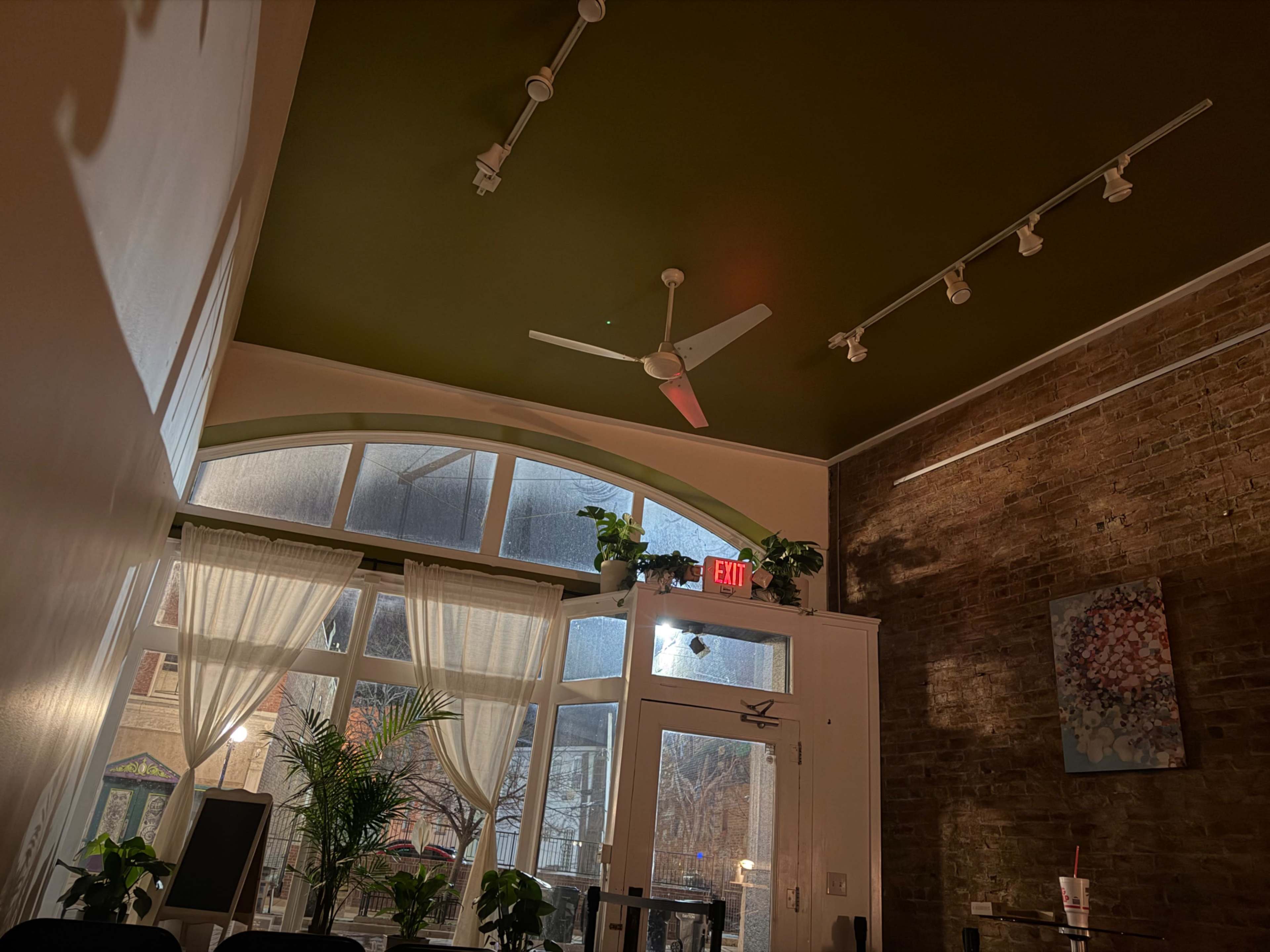 The image shows an interior space with a high ceiling, featuring a green ceiling, exposed brick walls, a ceiling fan, and large windows allowing natural light to enter.