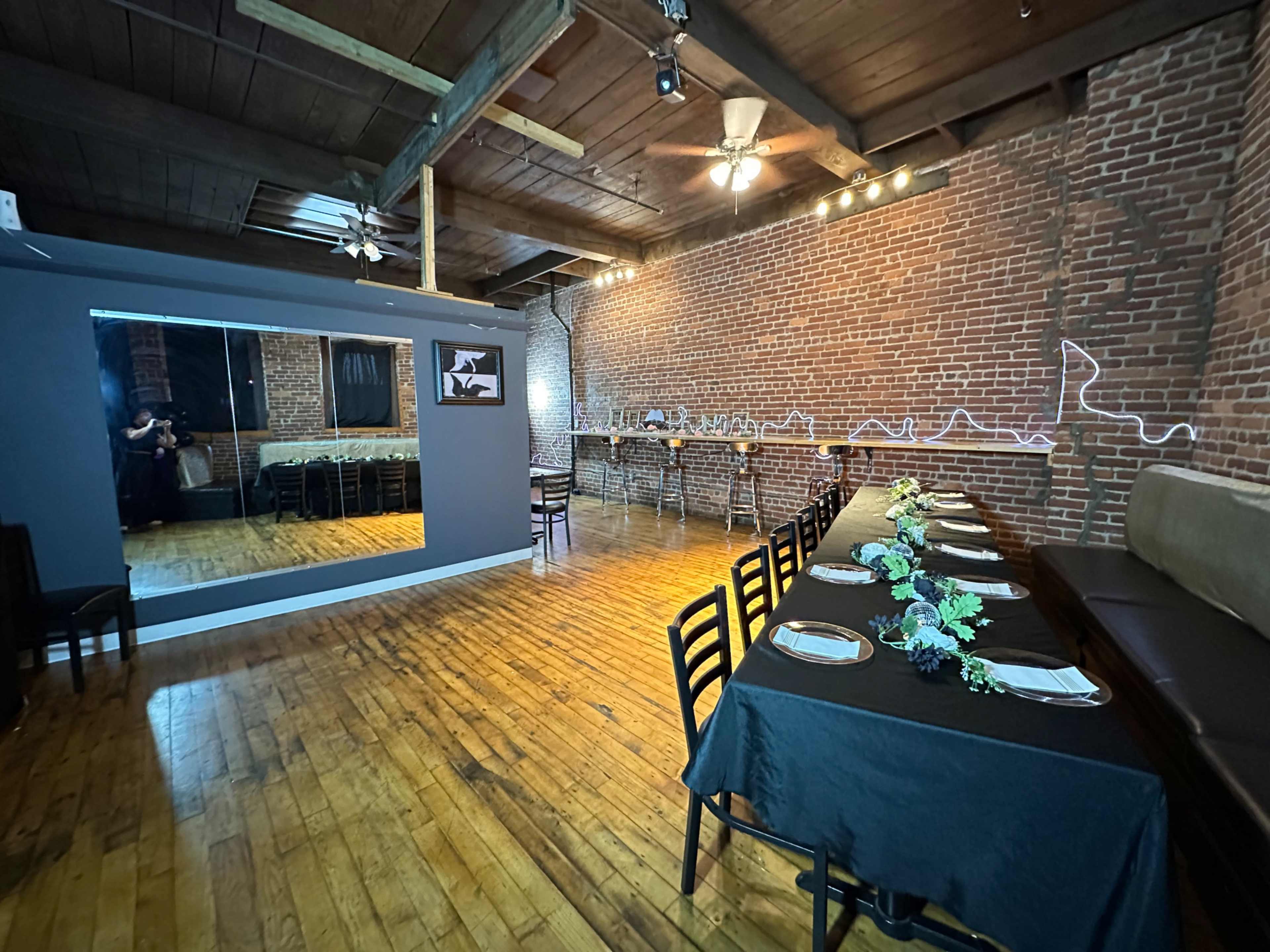 The image shows a spacious dining area with exposed brick walls, wooden floors, and a long table set for a meal, illuminated by wall-mounted lights.