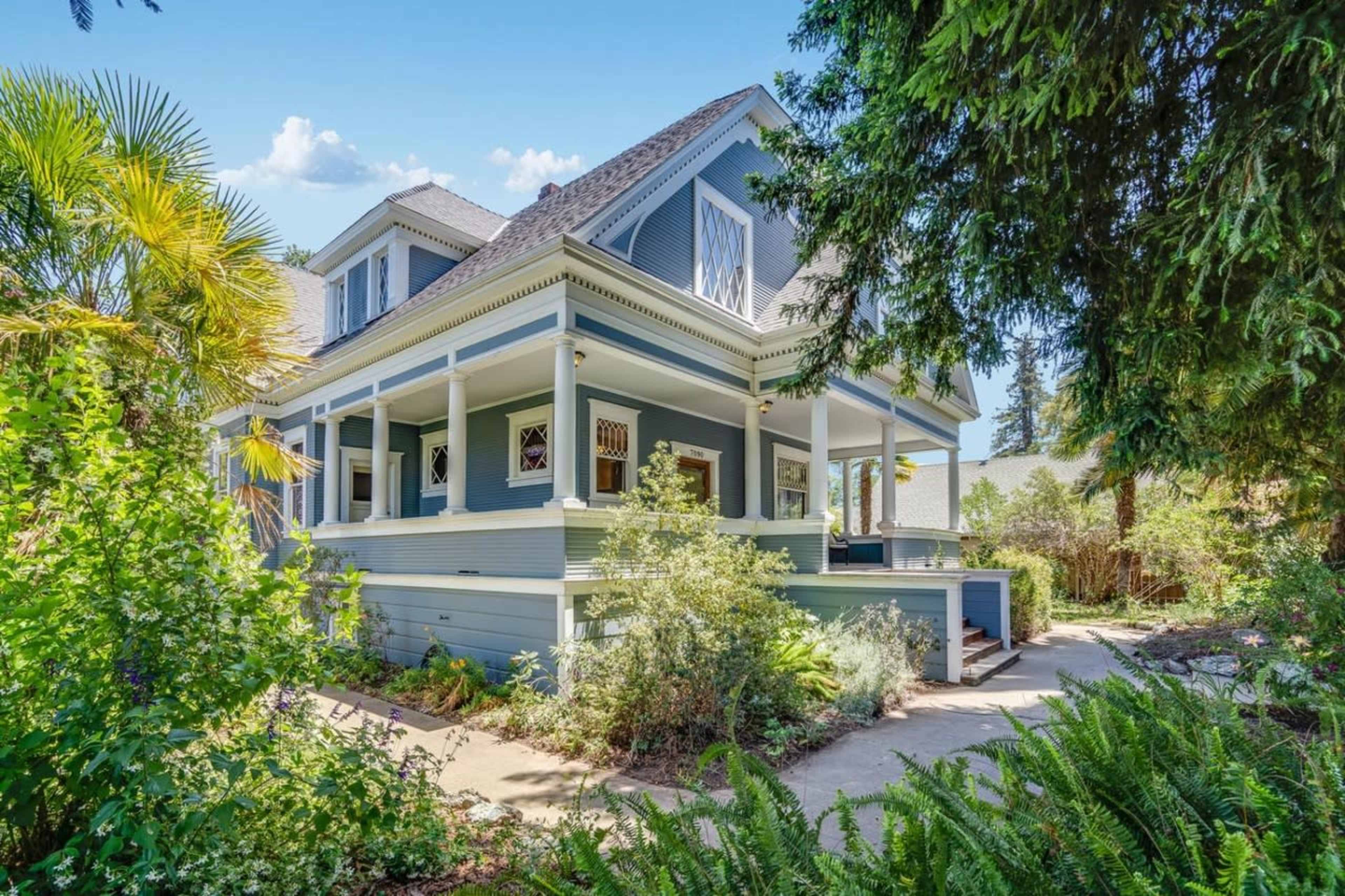 A large, two-story house with a blue exterior, surrounded by lush greenery and a well-maintained garden.