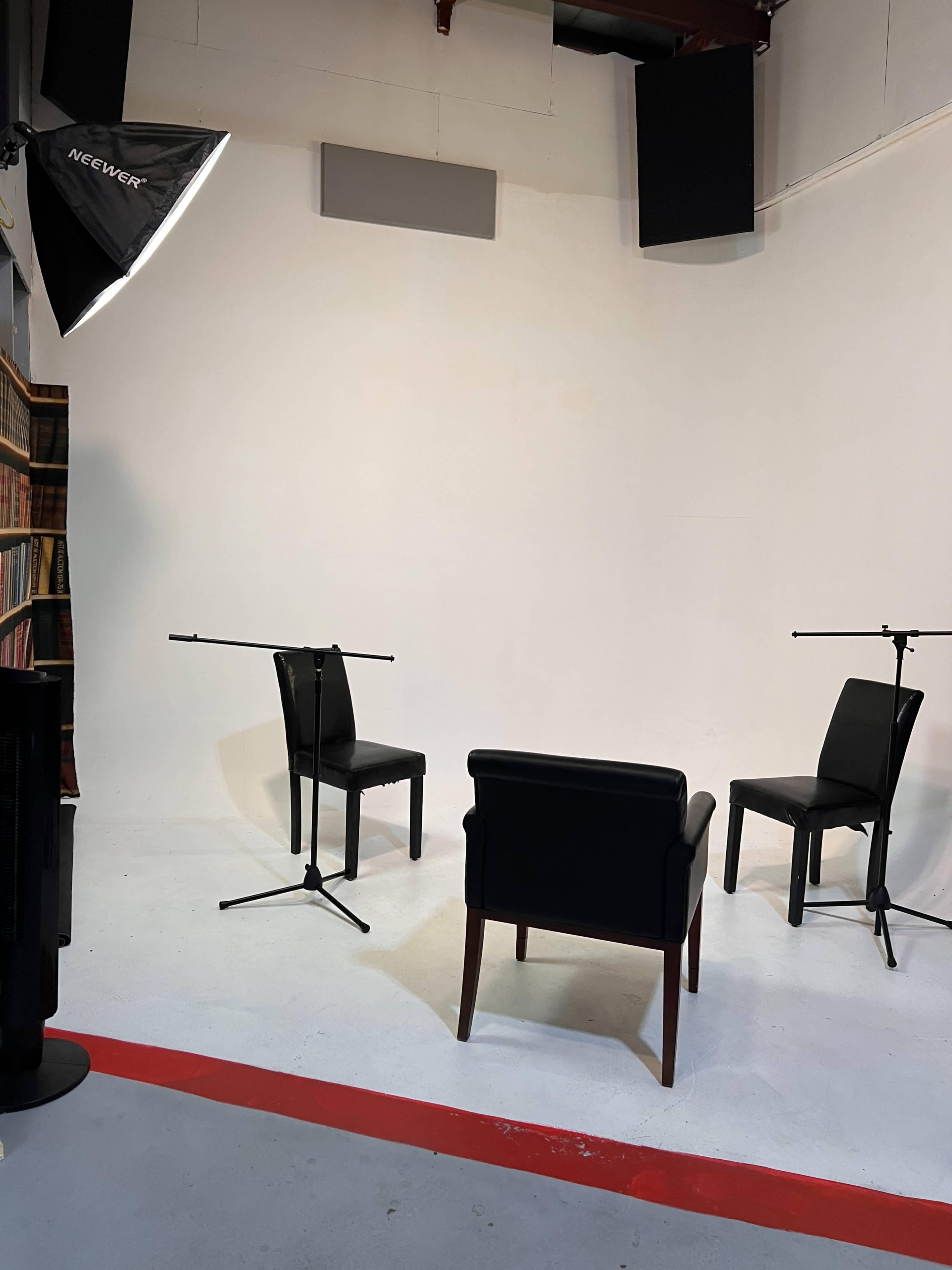 A minimalist studio setup with three chairs, two of which are black and one is a leather armchair, positioned in front of a plain white wall, with microphones on stands for recording.