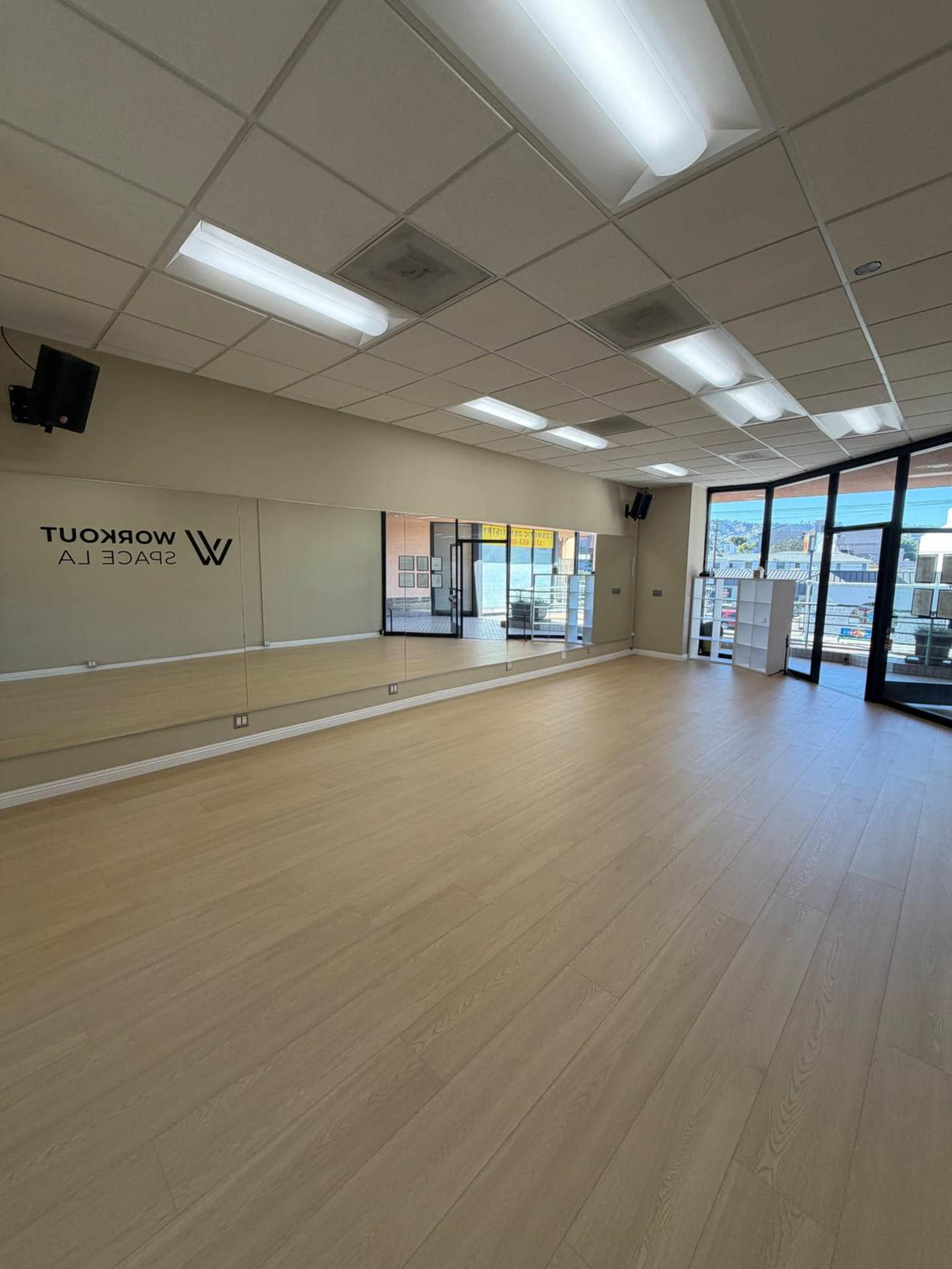The image shows a spacious workout studio with large mirrors, wooden flooring, and windows allowing natural light to enter.