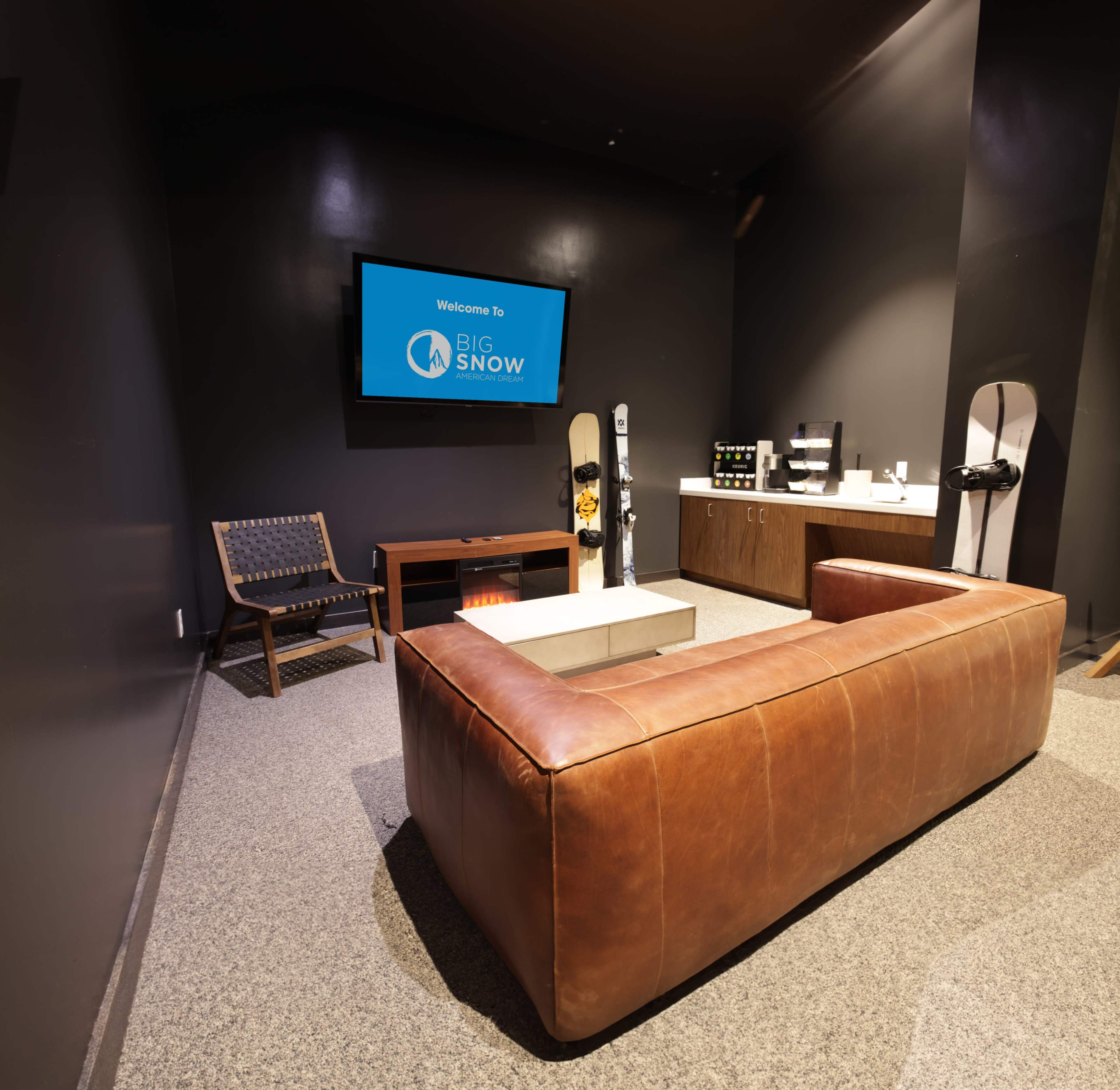 The image shows a cozy lounge area with a brown leather couch, a television displaying "Welcome to Big Snow," and winter sports equipment, including snowboards, against a dark wall.