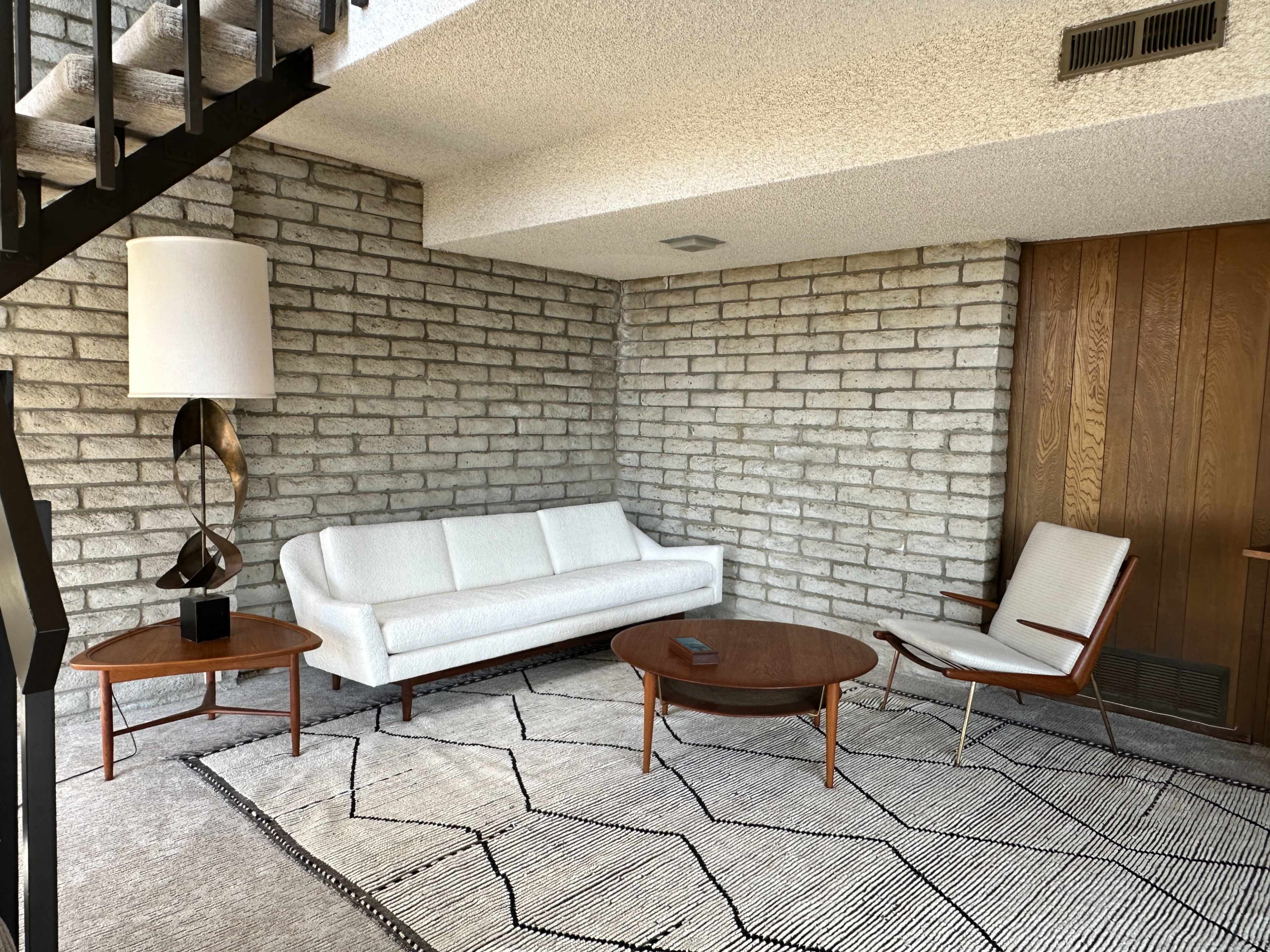 The image shows a minimalist living room with a white sofa, a round wooden coffee table, and an armchair, all set against a textured stone wall and a patterned rug.