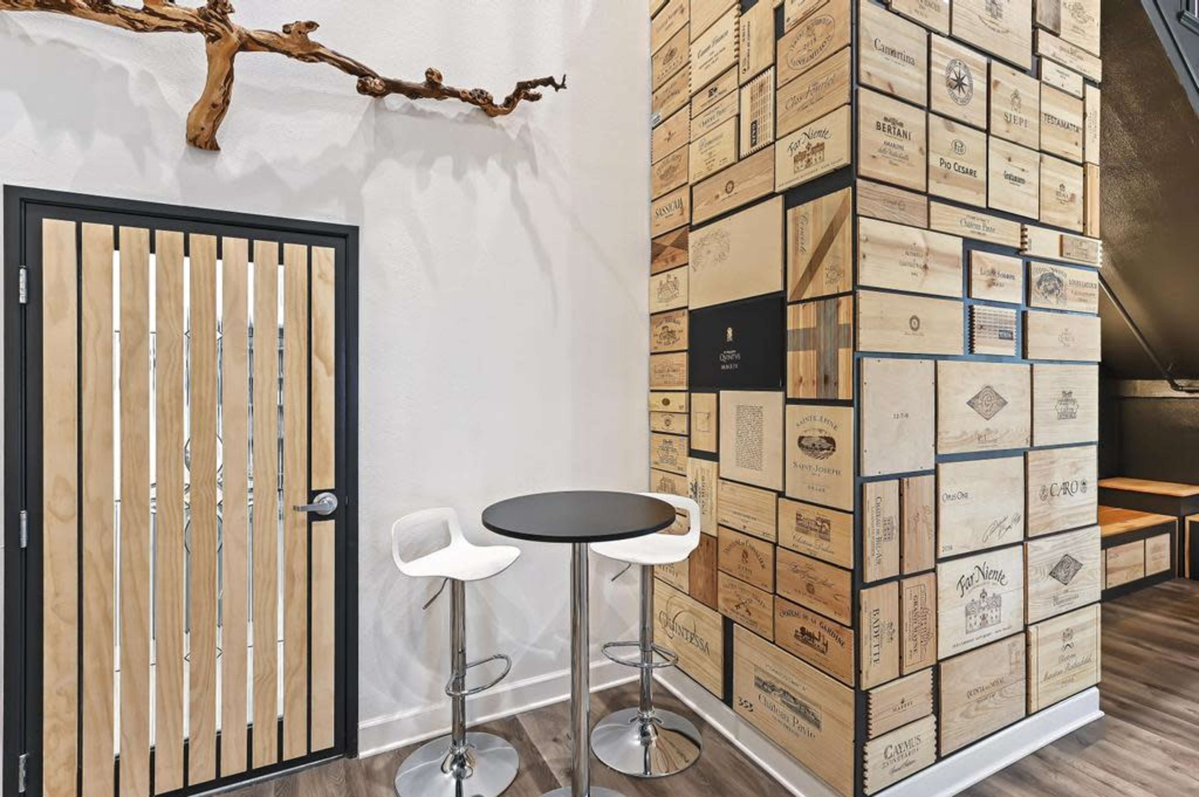 A modern interior space featuring a wooden door and a tall wall adorned with stacked wooden boxes, along with a round black table and two white stools in the corner.