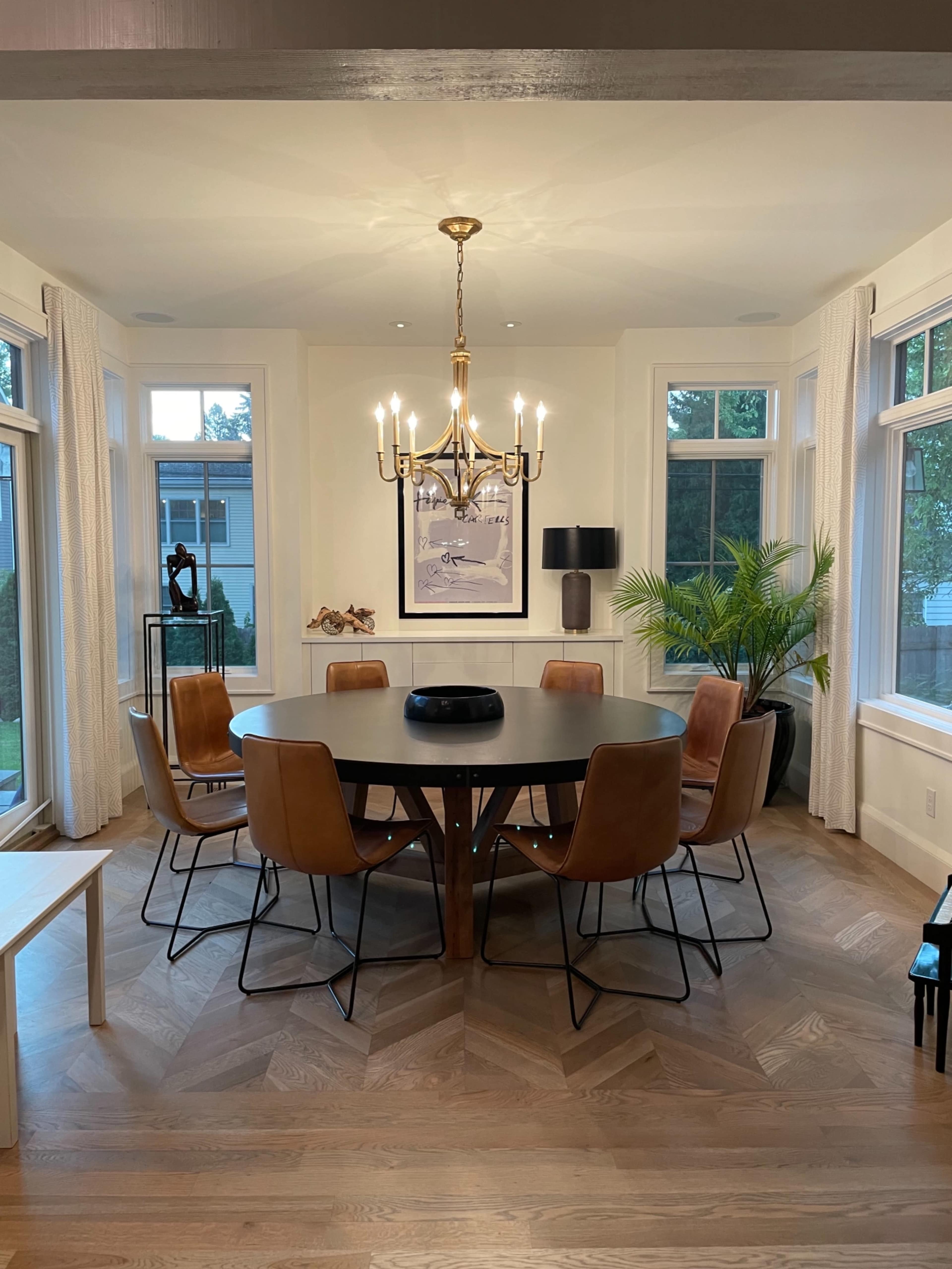A circular dining table surrounded by leather chairs is illuminated by a chandelier in a well-lit dining room.