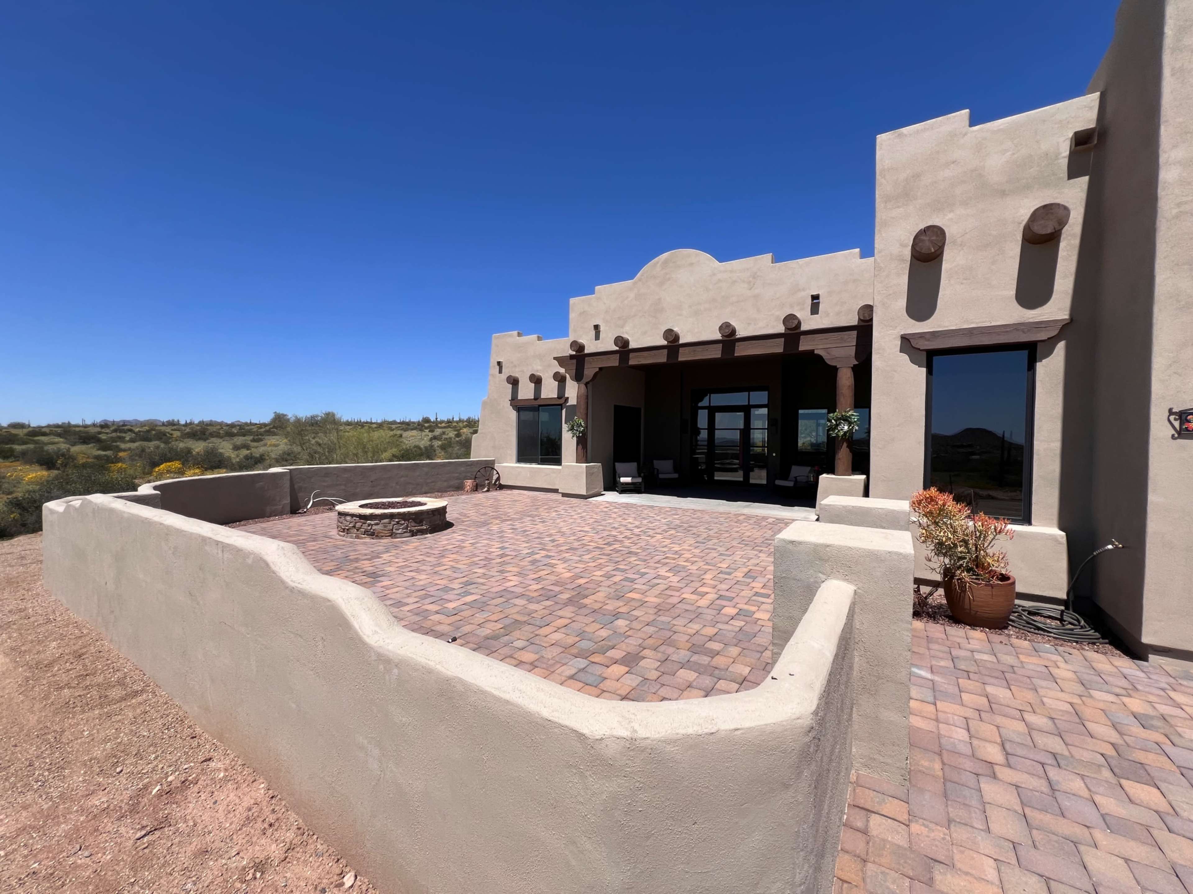 The image shows a southwestern-style home with a stucco exterior, a paved patio, and a stone fire pit surrounded by low walls.