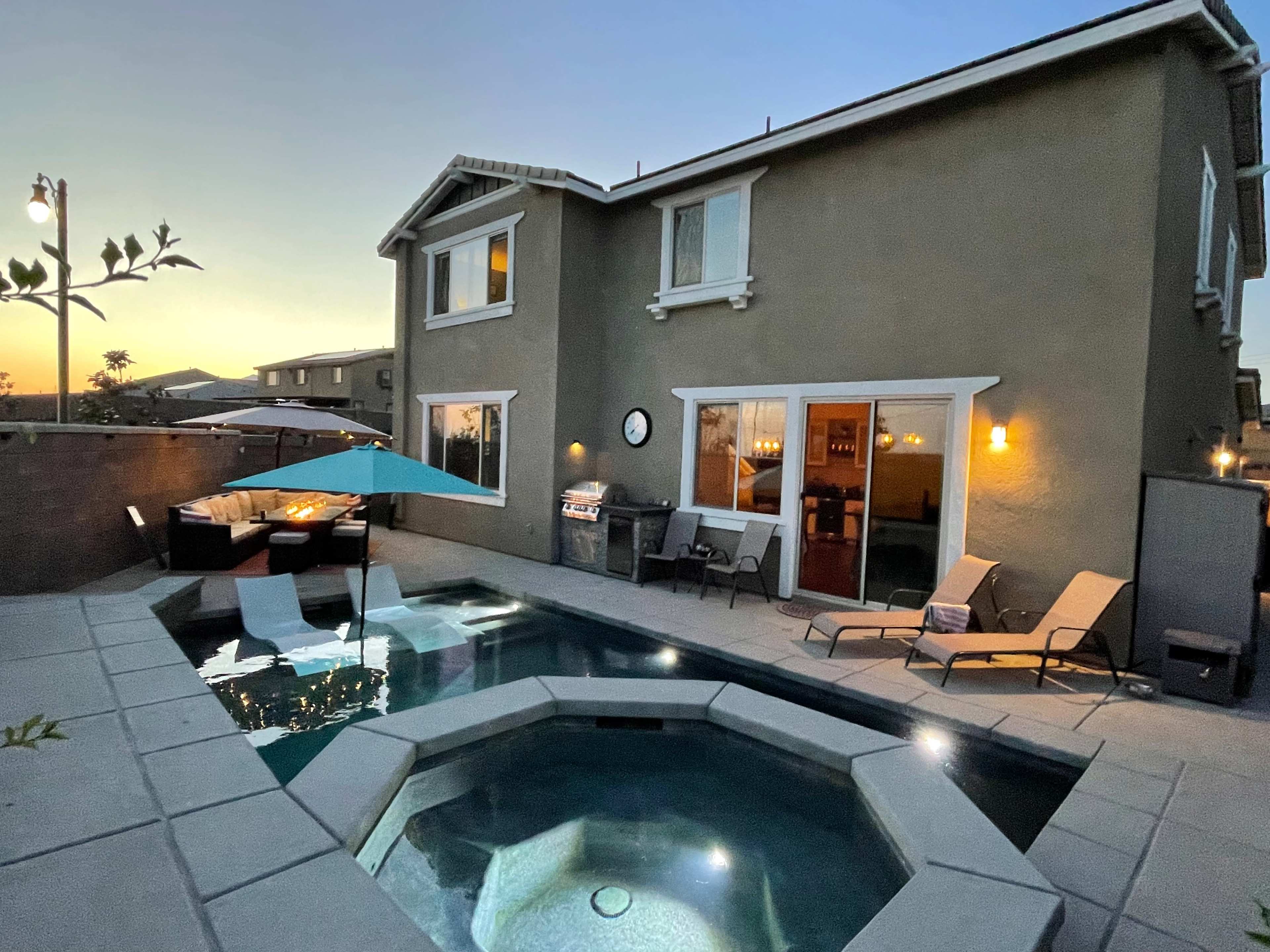 A modern home overlooking a pool area with lounge chairs, an umbrella, and a barbecue at dusk.