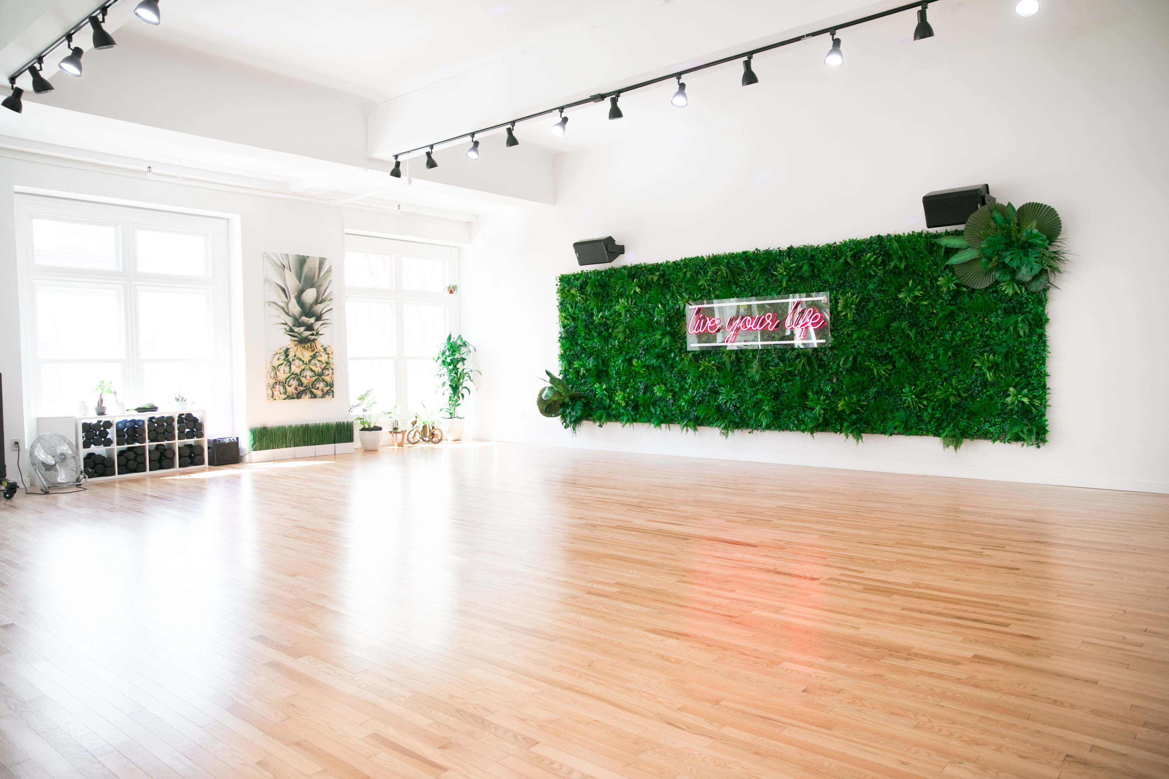 A spacious, well-lit indoor studio with wooden floors, a green plant wall featuring a neon sign, and two large windows allowing natural light to enter.